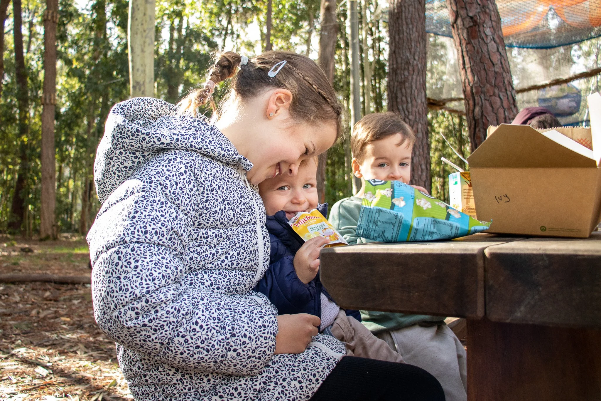 Three children sitting at a picnic table in a wooded area, with one holding a snack pouch and the others enjoying snacks, sunlight filtering through the trees.