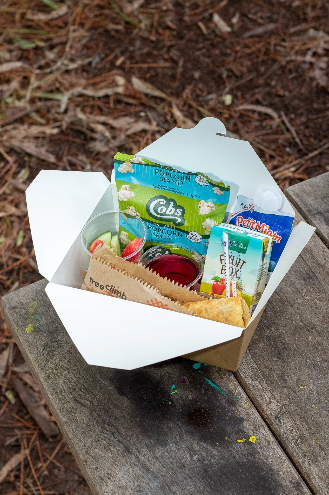 Open lunch box with popcorn, fruit drink, cut vegetables, and pastry on a wooden table.