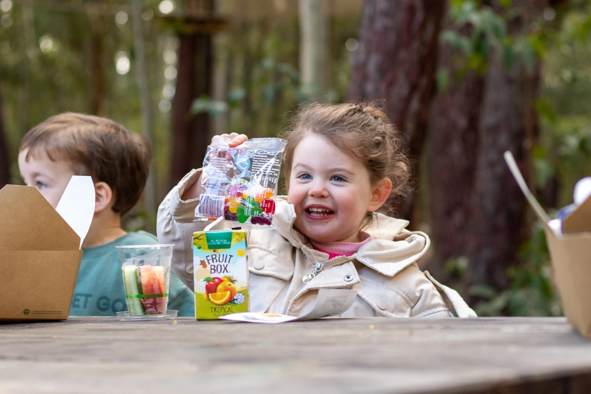 Two children at a picnic table, one holding a packet of gummy bears and a juice box, smiling. Another child is sitting next to a food container.