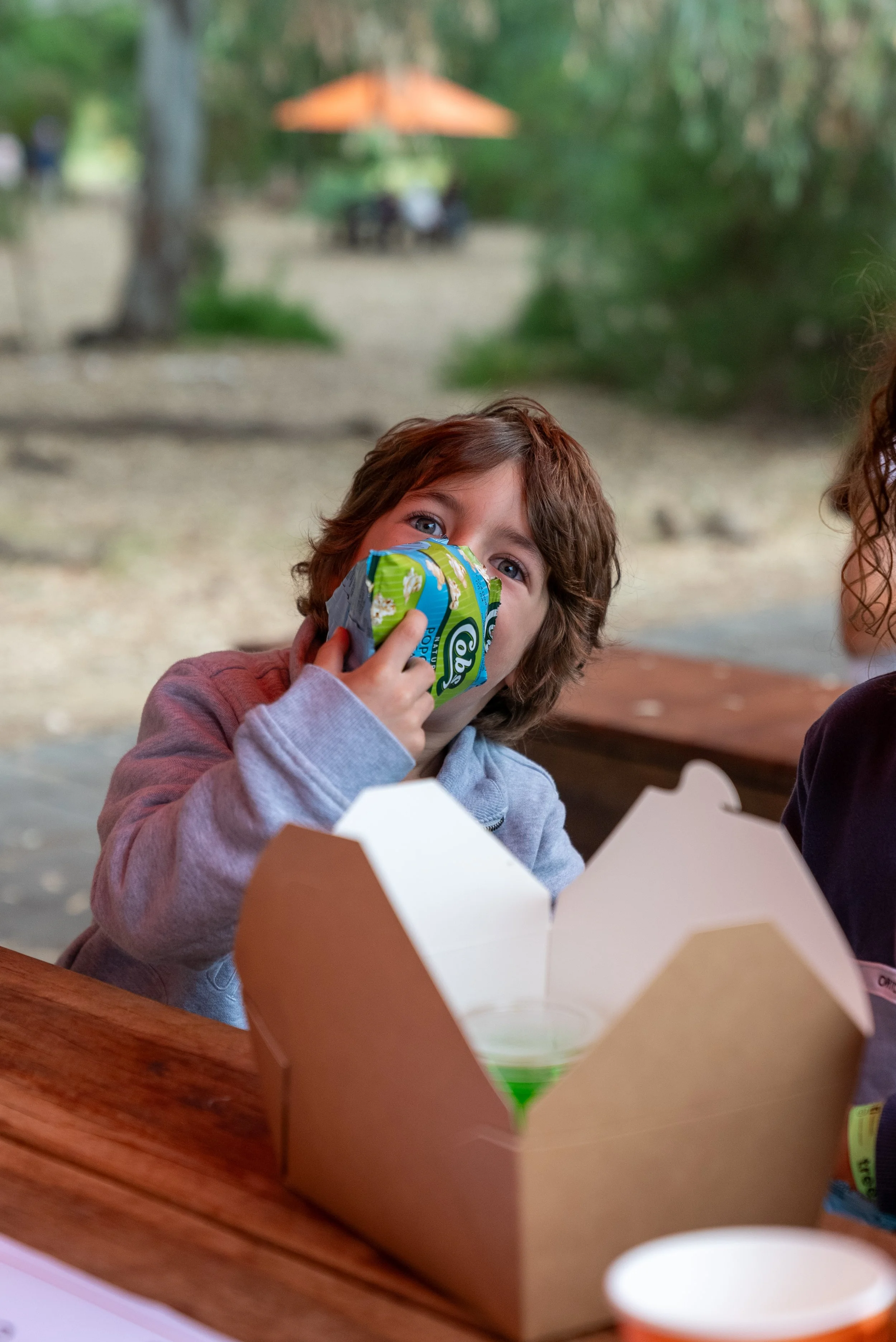 Child drinking from a juice pouch at an outdoor picnic table with a takeout box and cup nearby.