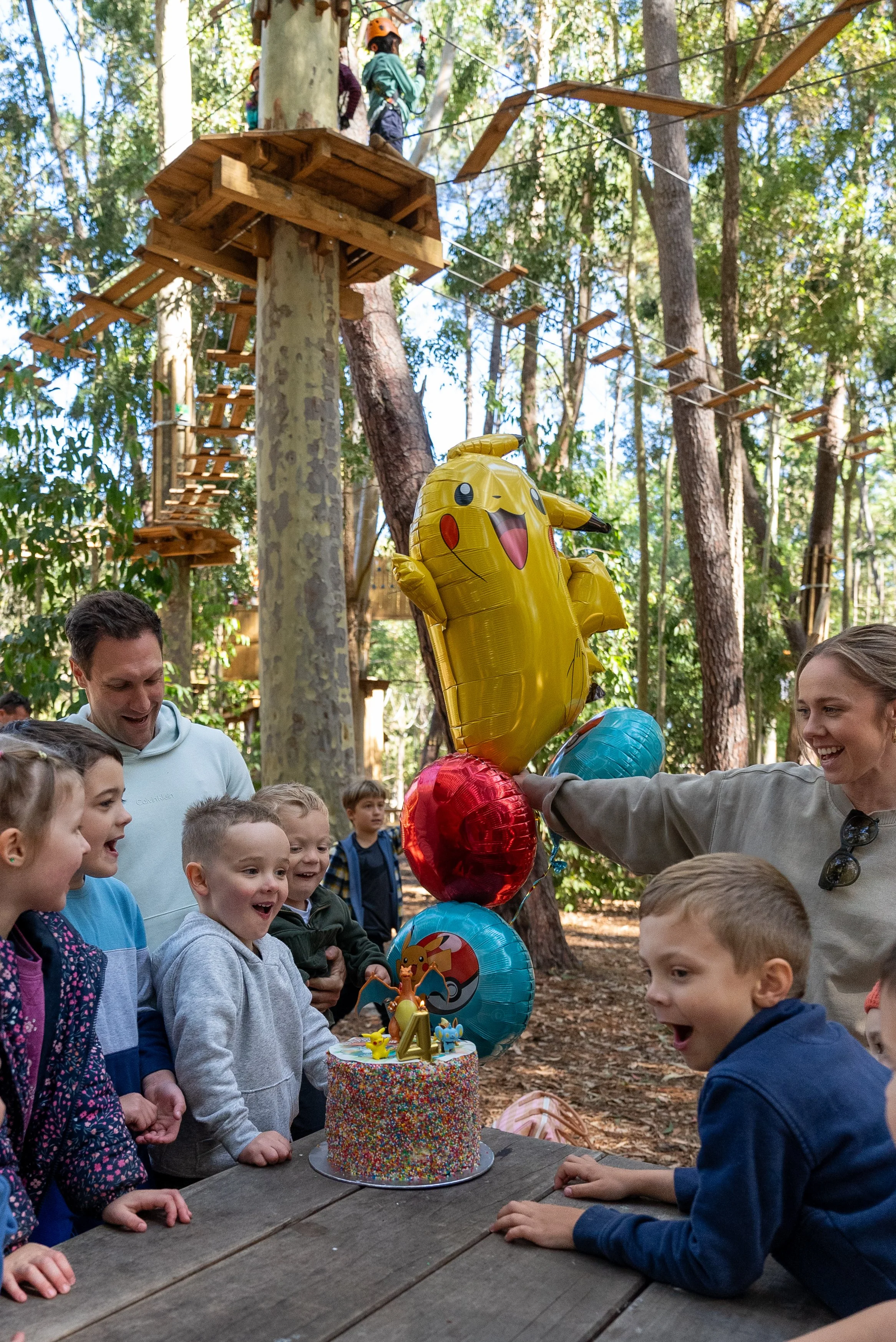 Children at a birthday party in a forest, gathered around a colorful cake with a Pikachu balloon.