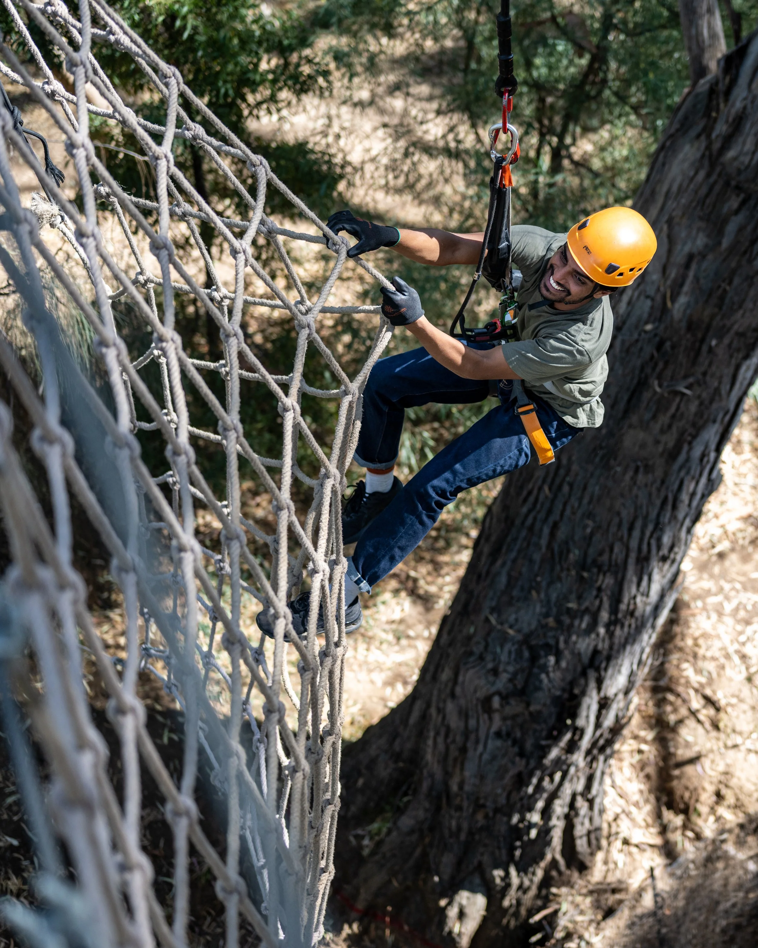 Person in an adventure park wearing a helmet and climbing a net obstacle in a forested area.