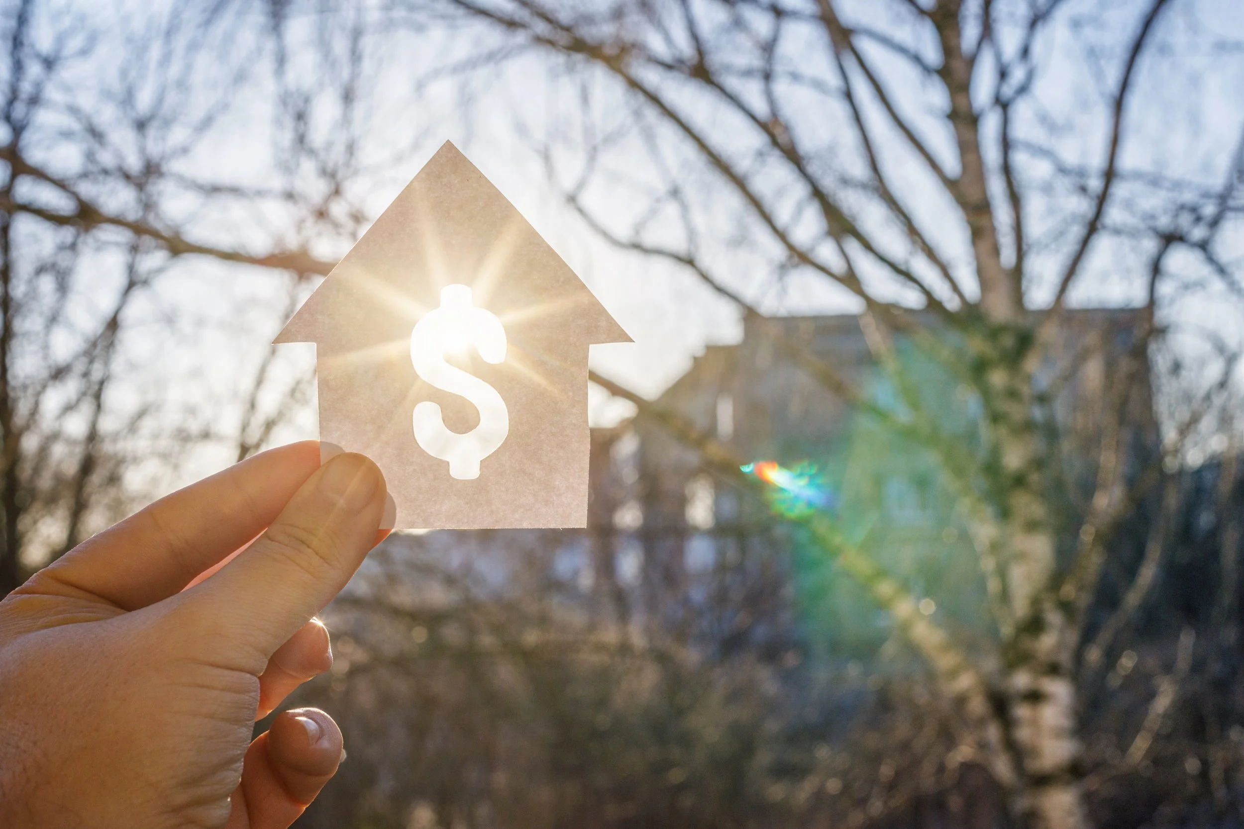 Hand holding a piece of paper with a dollar sign cutout, held up against the sun with a house-shaped arrow pointing upward. Background includes leafless trees and a building, with sunlight creating lens flare.