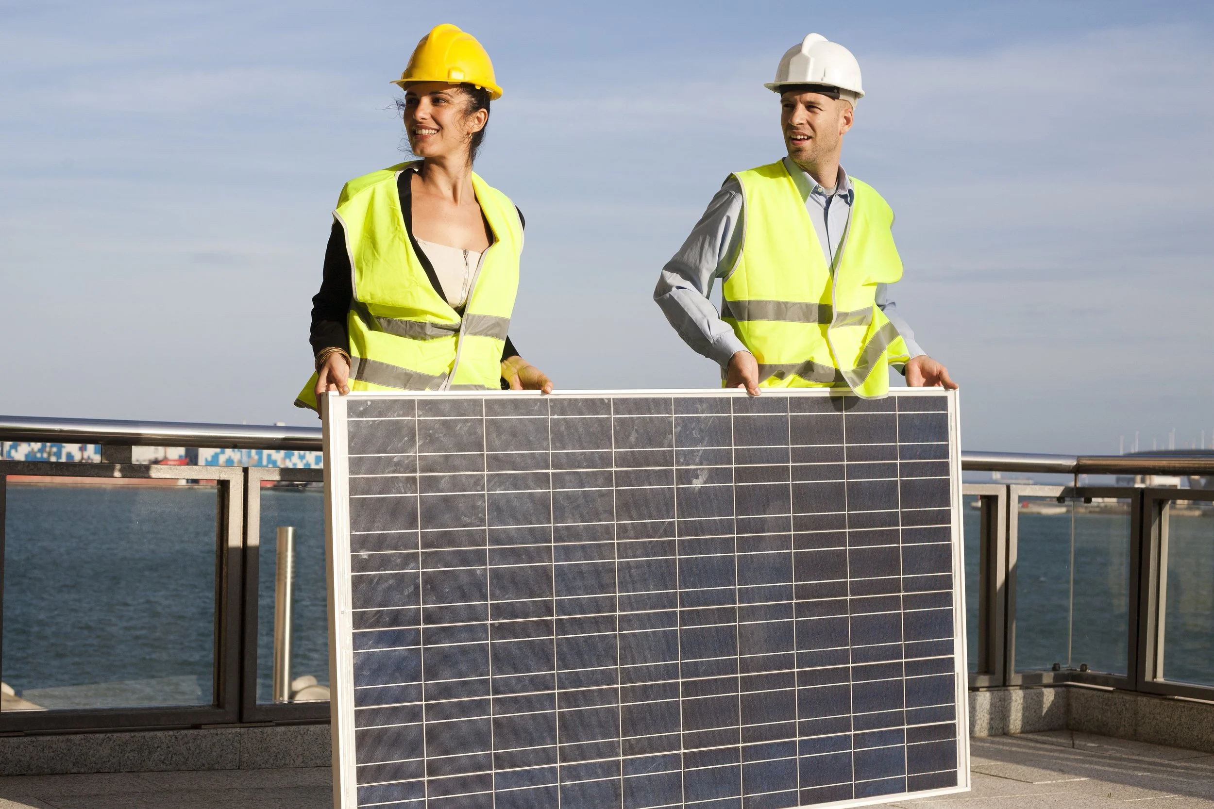 Two people in safety vests and helmets holding a solar panel outdoors near water with a cloudy sky.