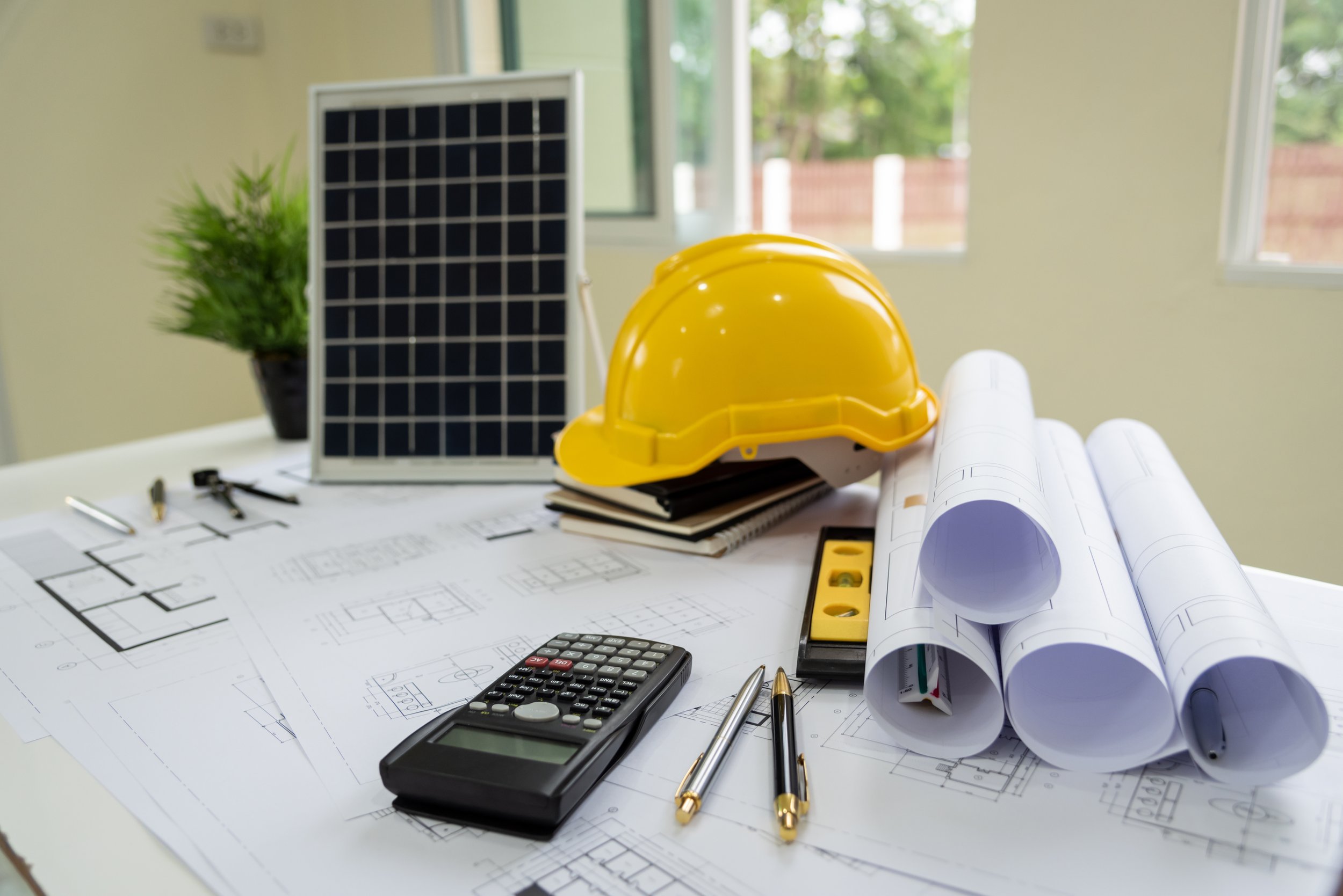 Architectural plans, a yellow safety helmet, blueprints, pens, a calculator, and a level on a white table in a bright room with windows.