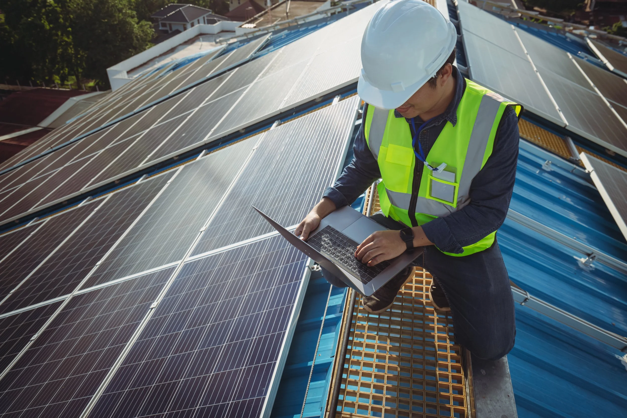 A technician wearing a white helmet and a fluorescent yellow safety vest working on solar panels on a rooftop using a laptop.