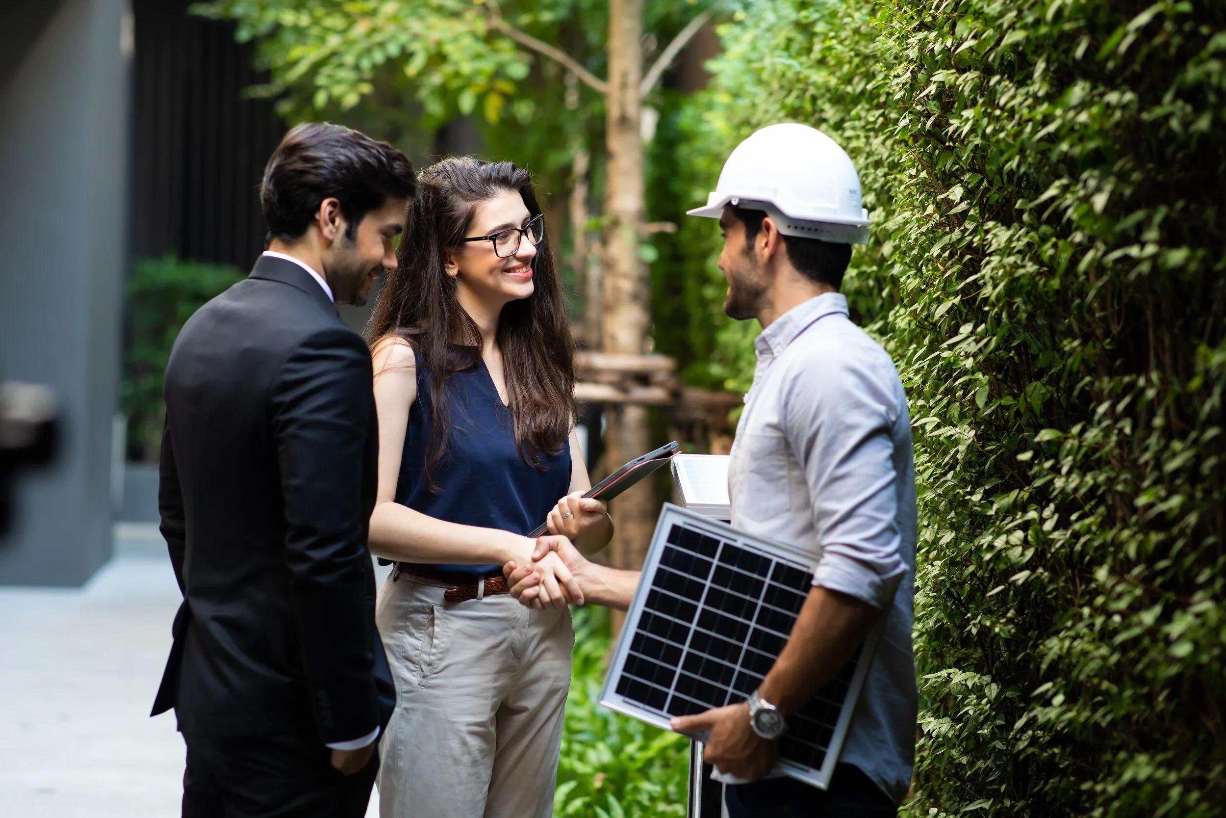 A woman and a man in a suit shaking hands with an engineer holding solar panels and a tablet, outdoors with greenery