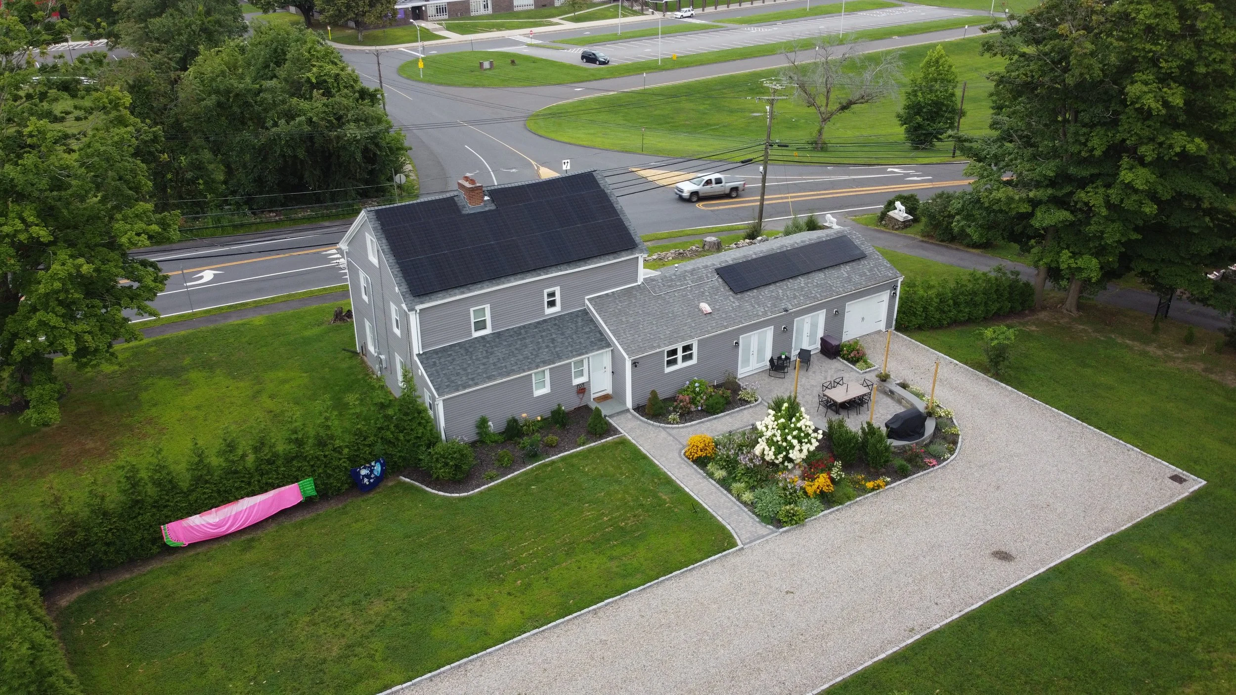 A wooden house with solar panels on the roof, surrounded by green trees and a landscaped yard with bushes and a driveway with a parked black SUV.