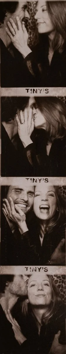 A strip of four black-and-white photo booth pictures featuring a man and a woman. In the first image, the woman is smiling and looking at the man, touching his face. In the second, they are close with the woman leaning in, her hand touching the man's face. In the third, they are smiling widely, with the woman holding her hand to the man's cheek. In the last, the man is kissing the woman's cheek while she looks directly at the camera with a confident expression.