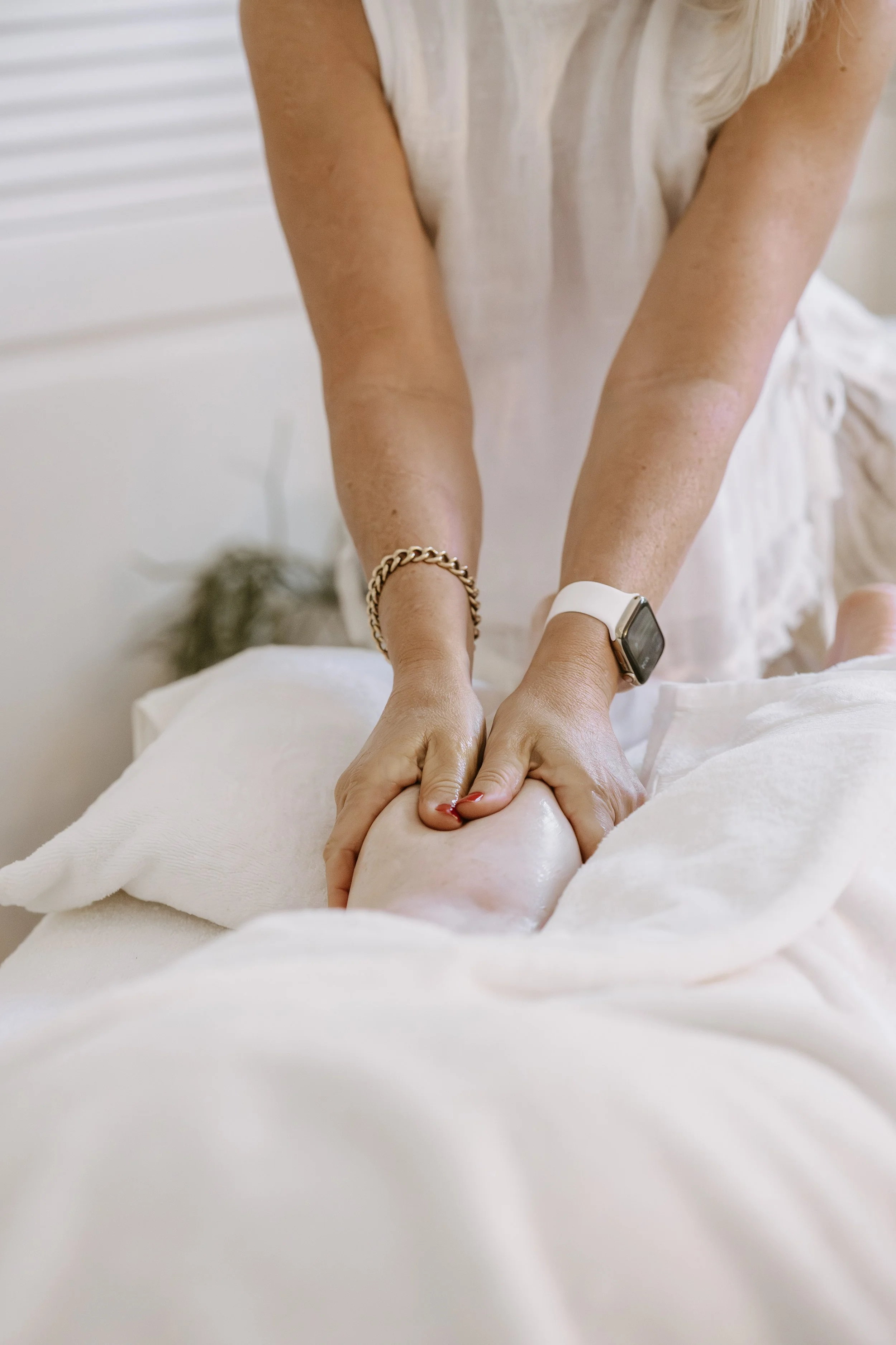 A woman sitting on a bed with white sheets, hugging her knees. She is wearing a white tank top. The logo in the bottom right corner reads 'COCO Massage & Beauty'.