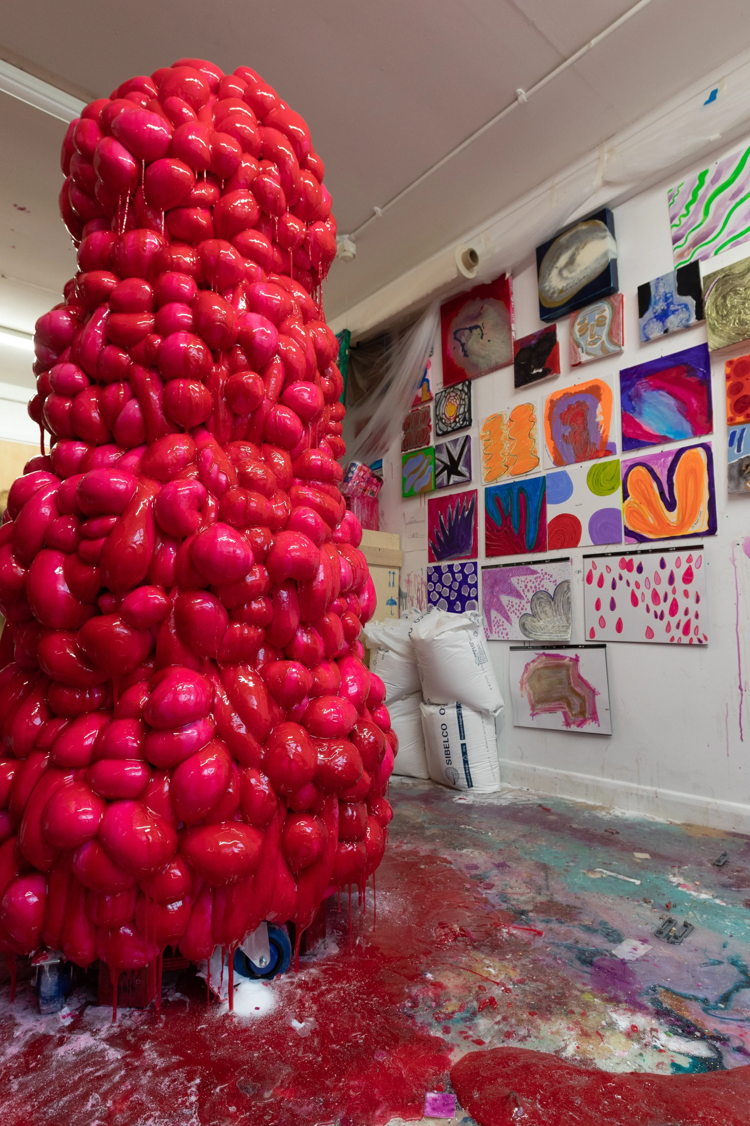 Large red sculpture made of shiny, rounded objects resembles a coral or organic form, located in an art studio with colorful paintings and mixed media artworks on the walls.