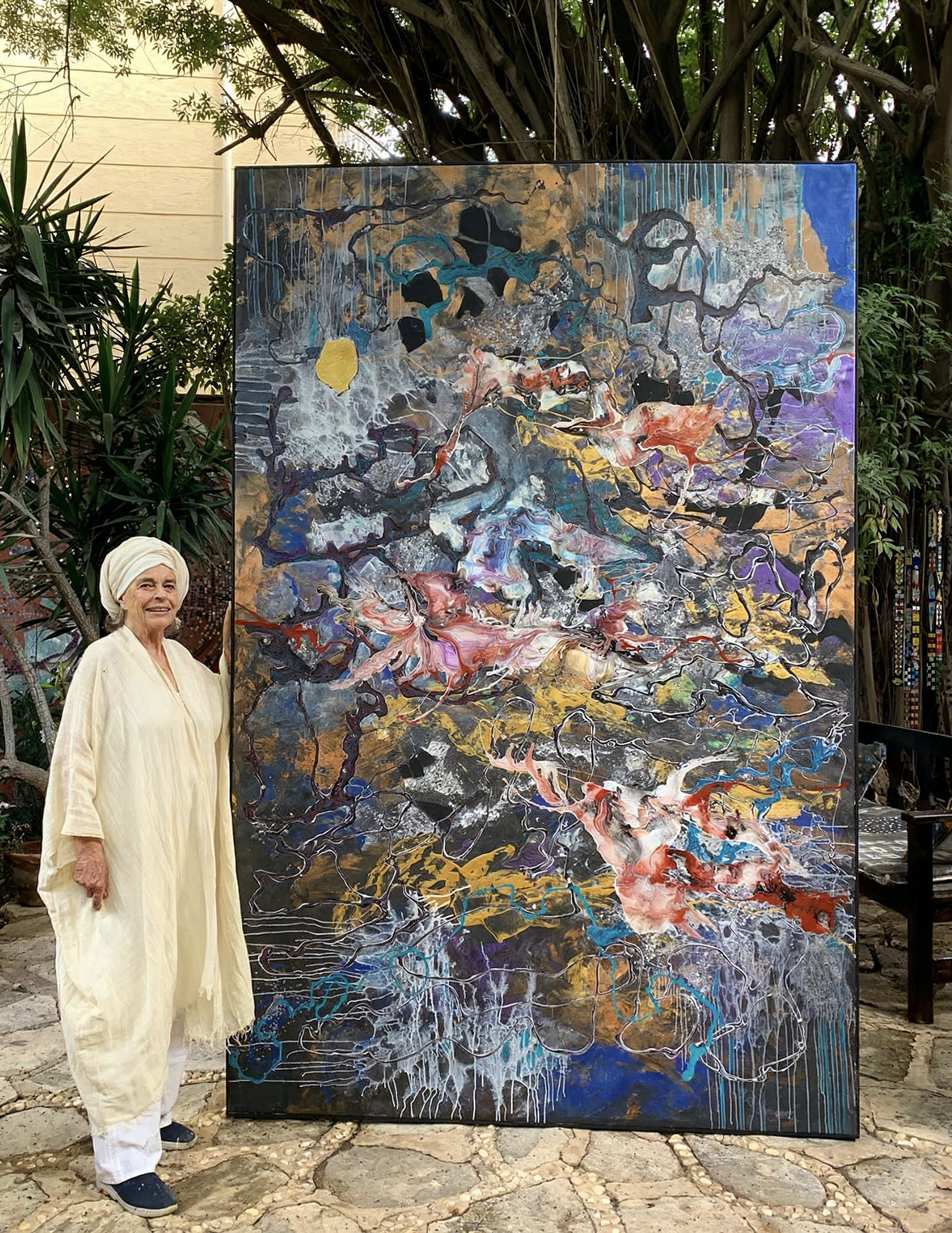 Older woman in light-colored clothing and headscarf standing next to a large abstract painting with swirling colors and textures, outdoors surrounded by greenery.