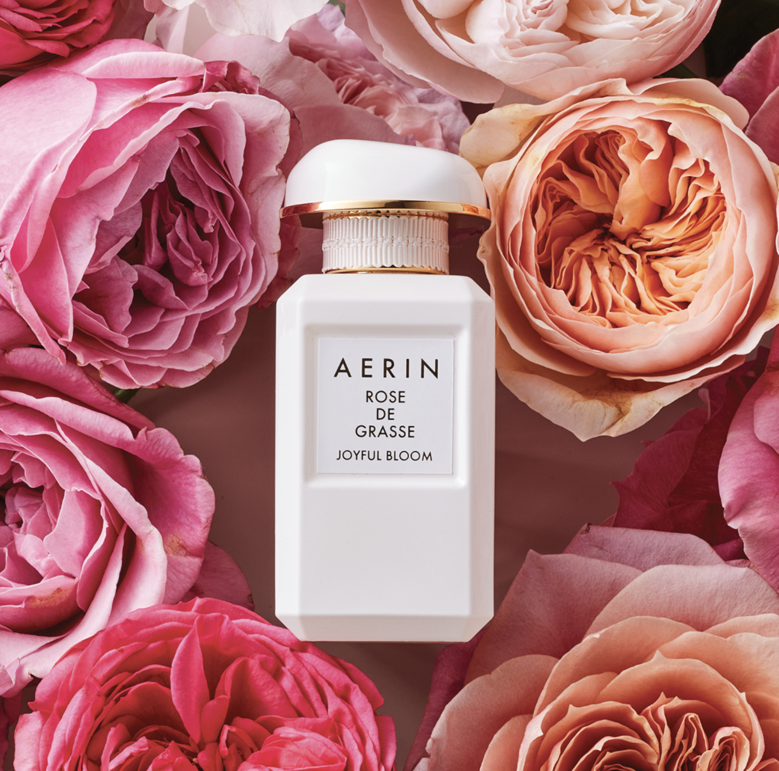 A white bottle of AERIN Rose de Grasse Joyful Bloom perfume surrounded by pink and peach roses.