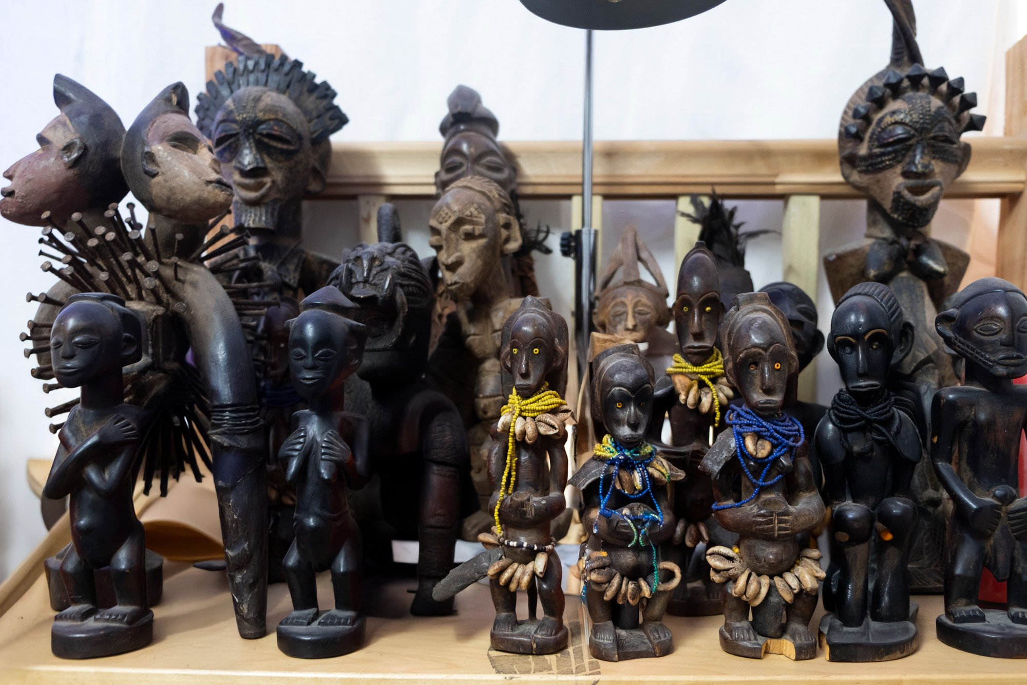 Display of various wooden tribal masks and statues, some adorned with colorful beads, arranged on a wooden shelf.