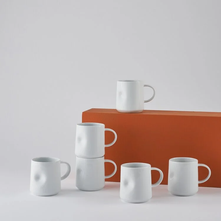 Six plain white ceramic coffee mugs arranged around a brown rectangular block, with one mug placed on top of the block, against a neutral background.