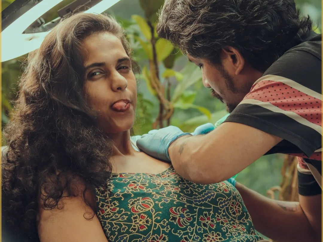 A woman sticking out her tongue while getting a tattoo on her upper arm from a man in a colorful shirt, with green foliage in the background.