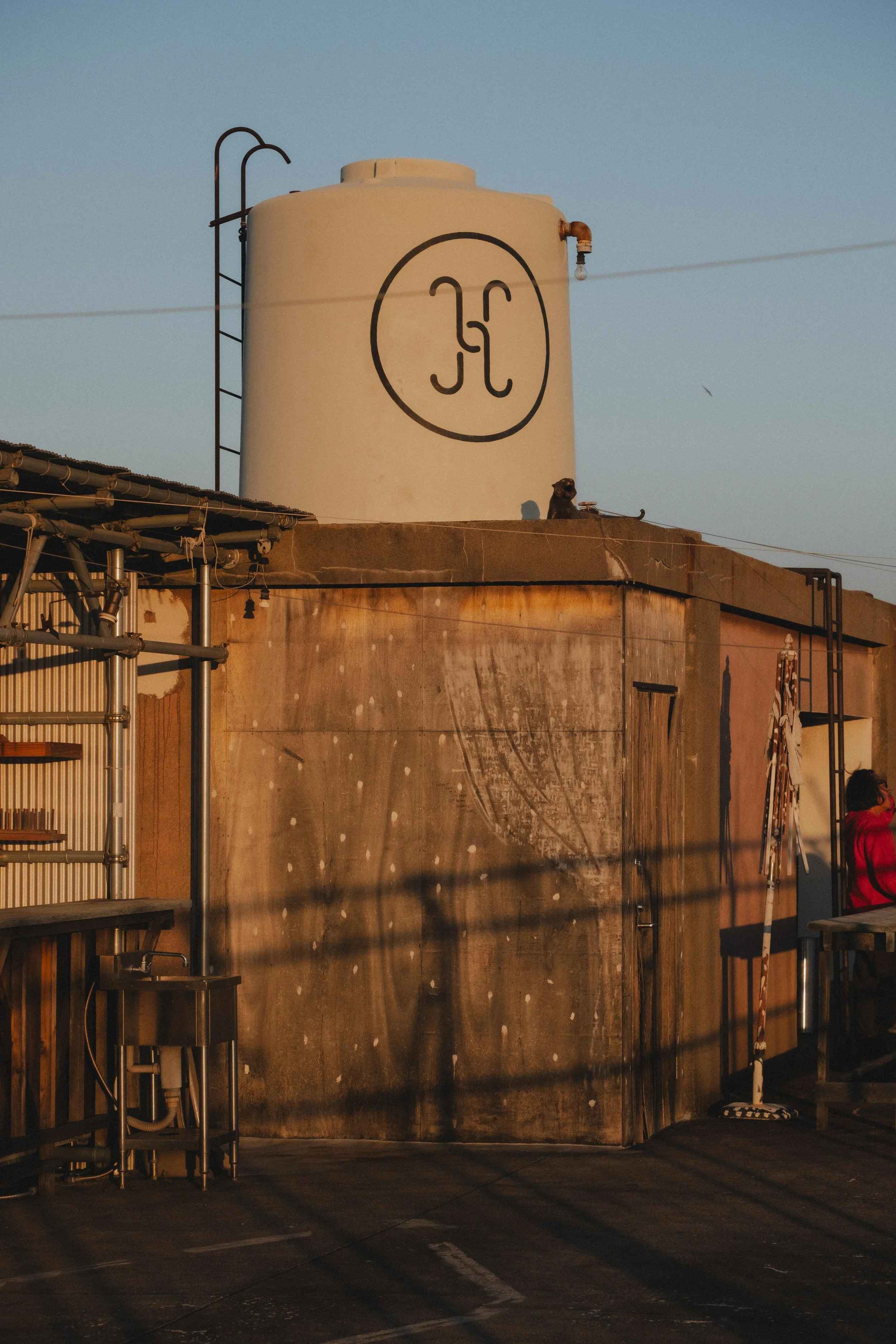 Industrial rooftop with a large white water tank featuring a circular logo with stylized 'H' and a dog sitting next to the tank, a ladder attached to the tank, and shadows cast on a rusty wall during sunset.