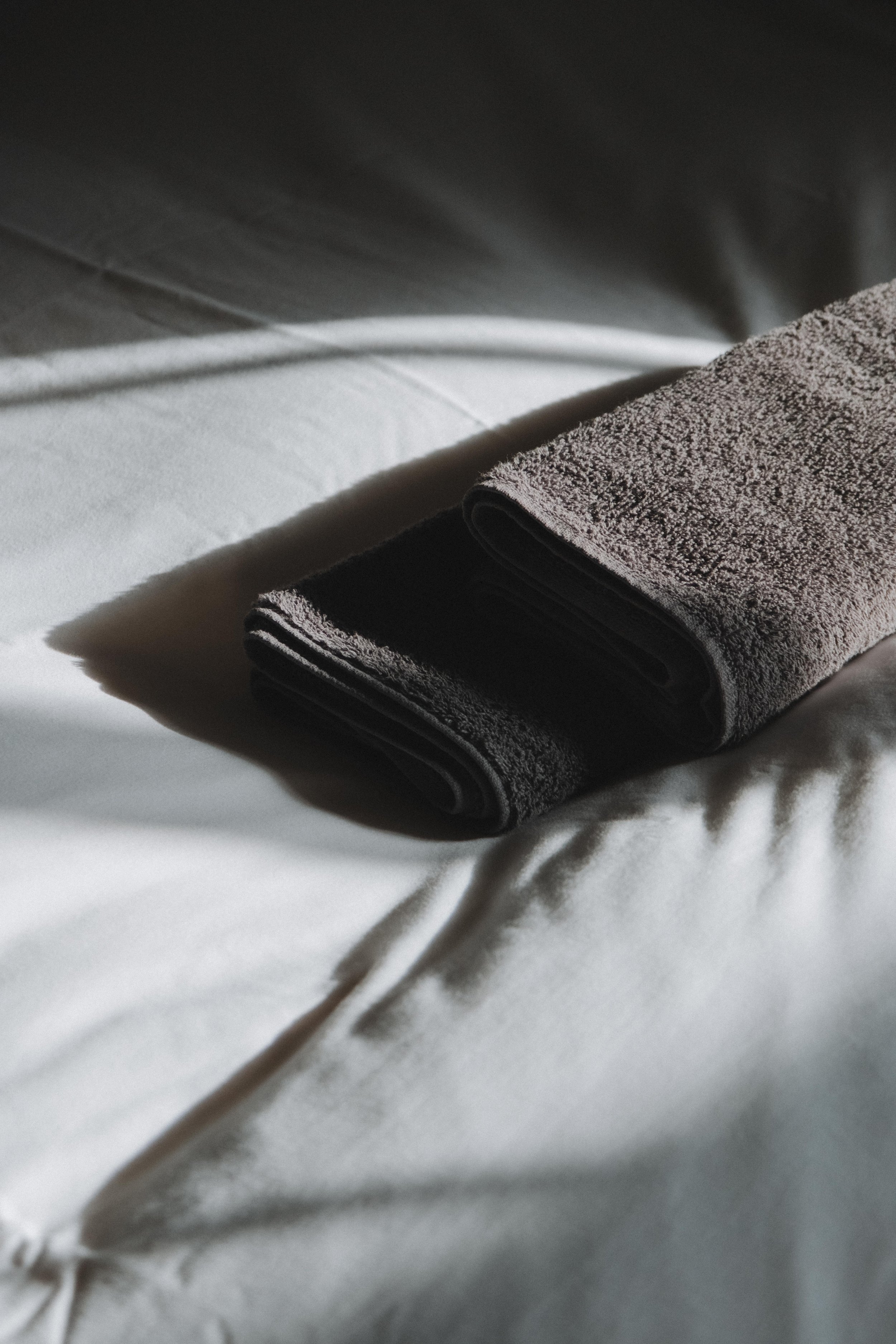 A rolled gray towel placed on a white bedsheet with soft lighting creating shadows.