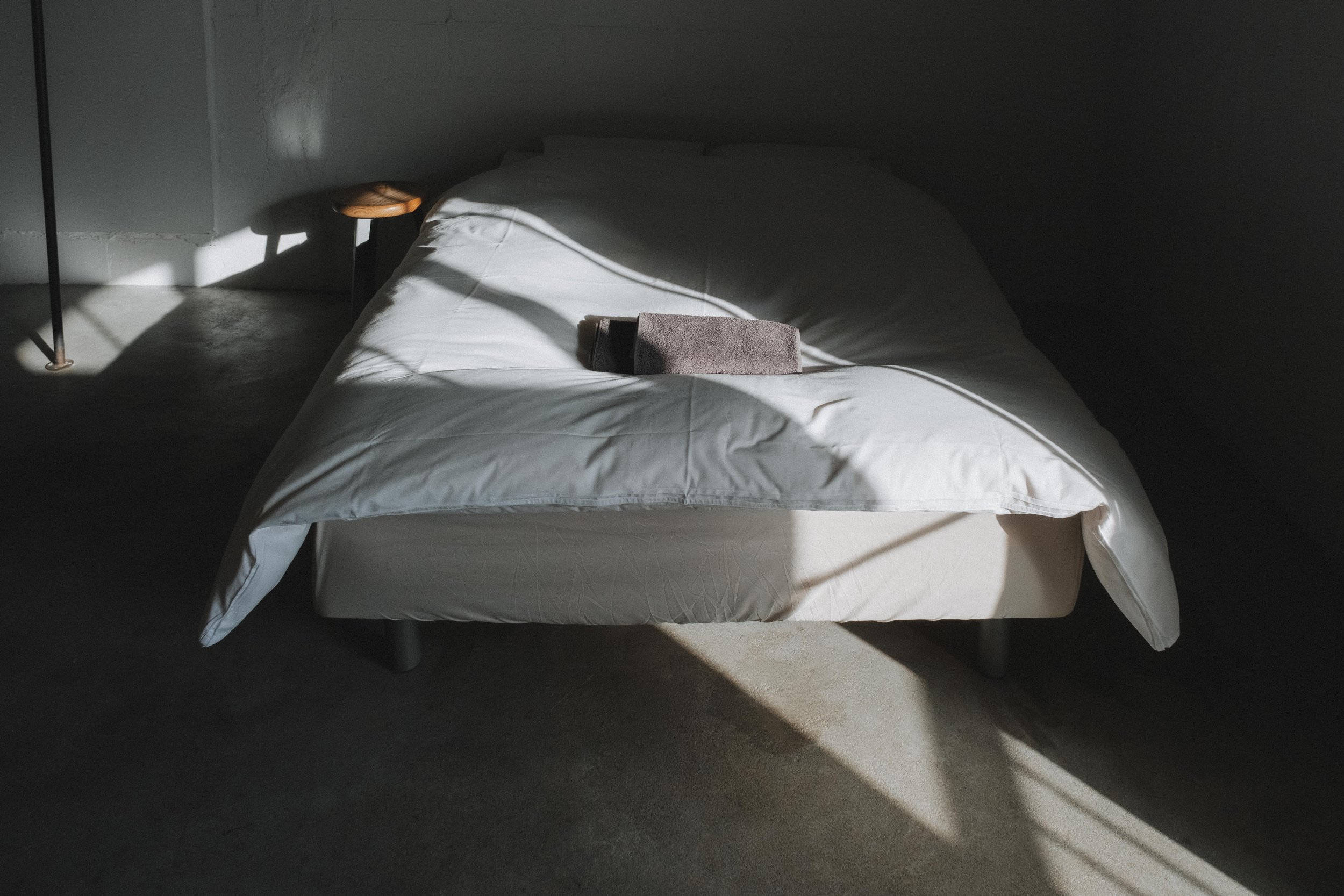 A neatly made bed with white sheets and a folded gray towel on top, illuminated by natural light in a minimalist bedroom.
