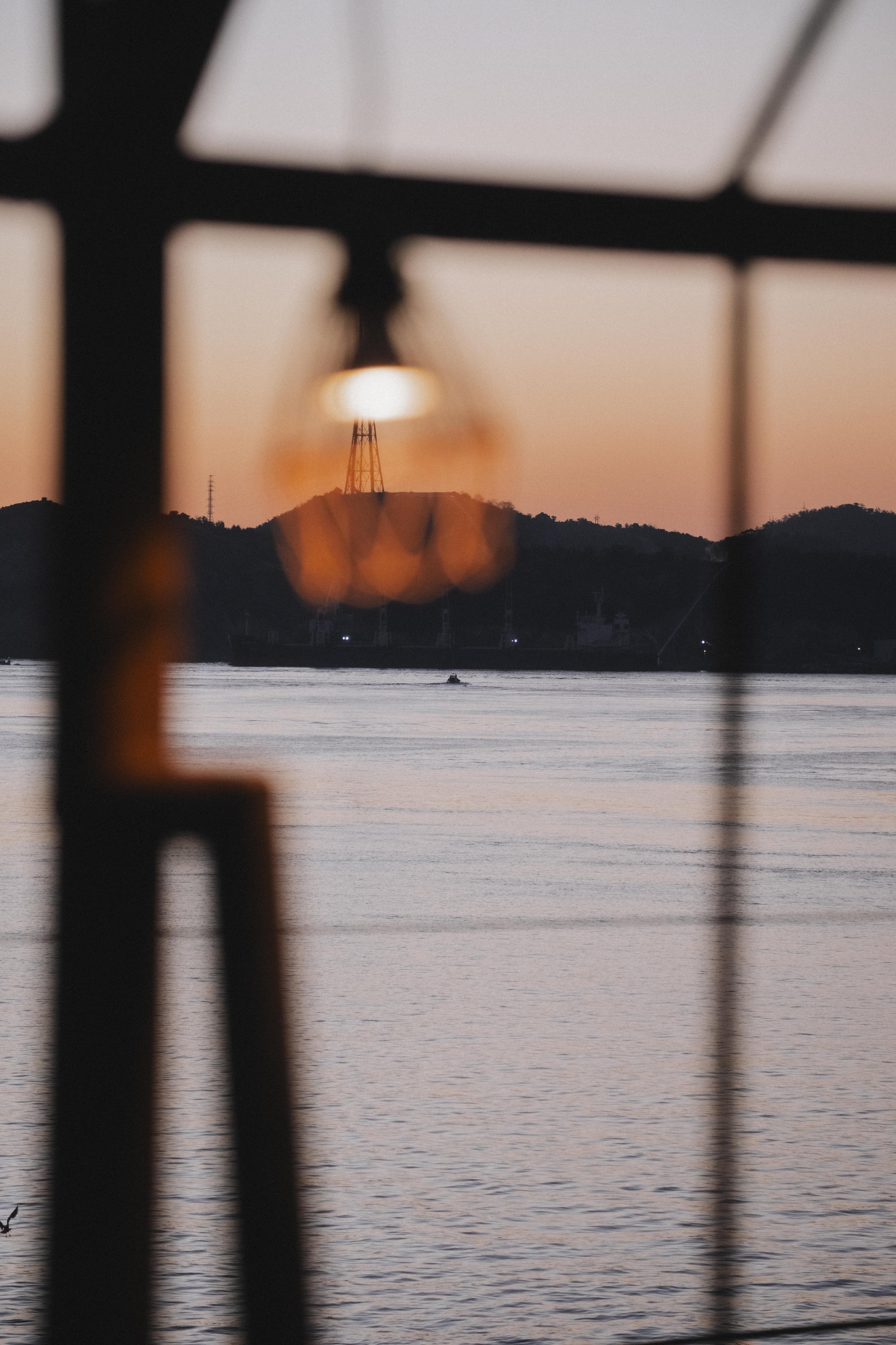 Sunset view over water framed by a structure, with hills and a transmission tower in the distance.