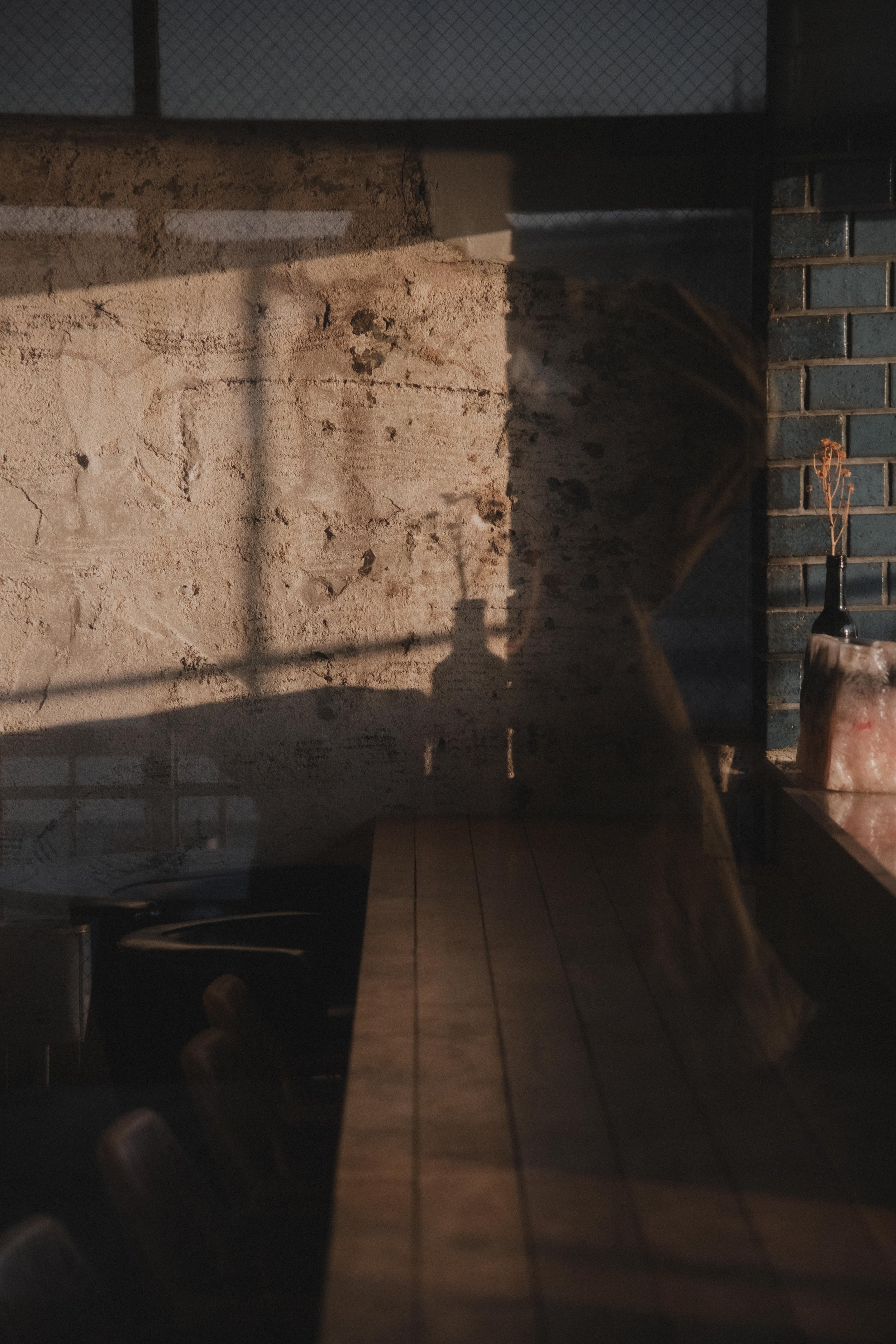 Shadow of a person taking a photograph reflected on a glass surface with an exposed brick wall, a blue tiled wall, and a wooden table inside a dimly lit room.