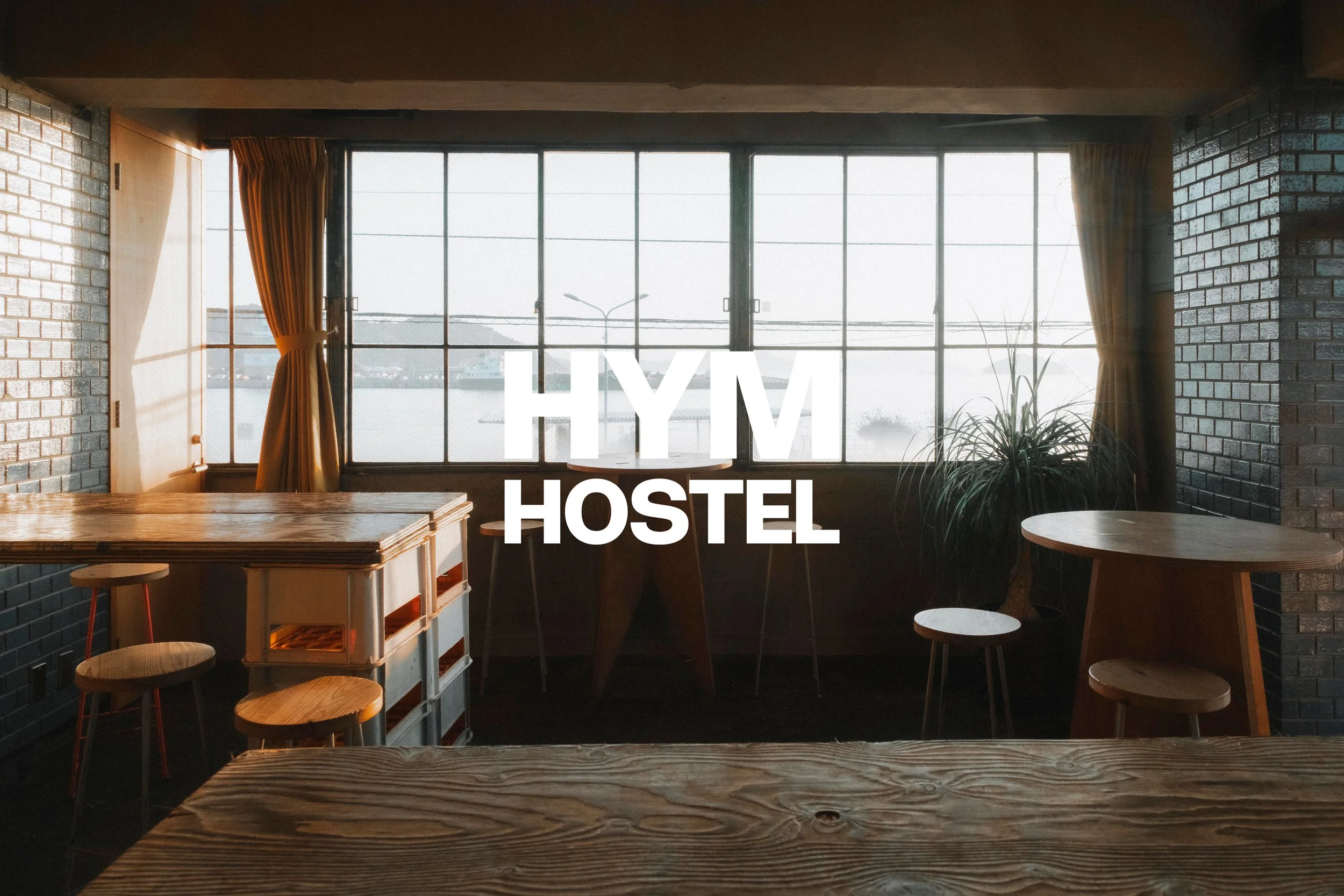 Interior of a rustic hostel with wooden tables, stools, and large windows with curtains, overlooking a river or lake.