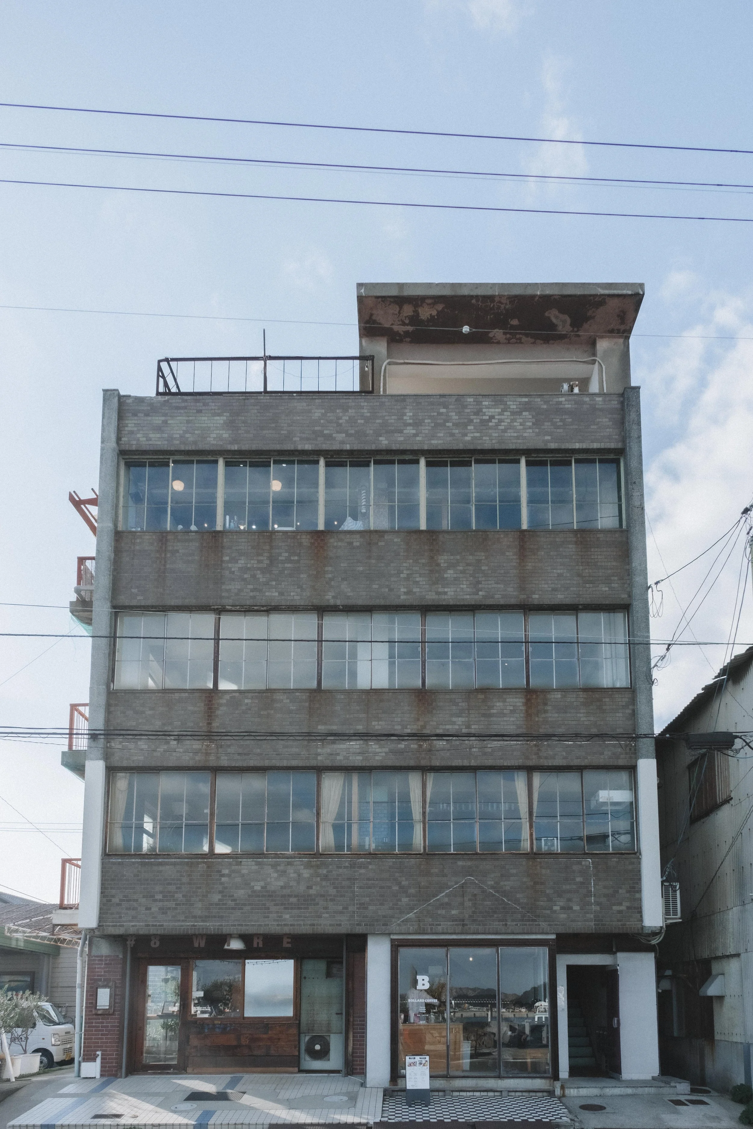 A multi-story building with large glass windows and a brown brick facade, with a small entrance at the bottom and balconies on the side.