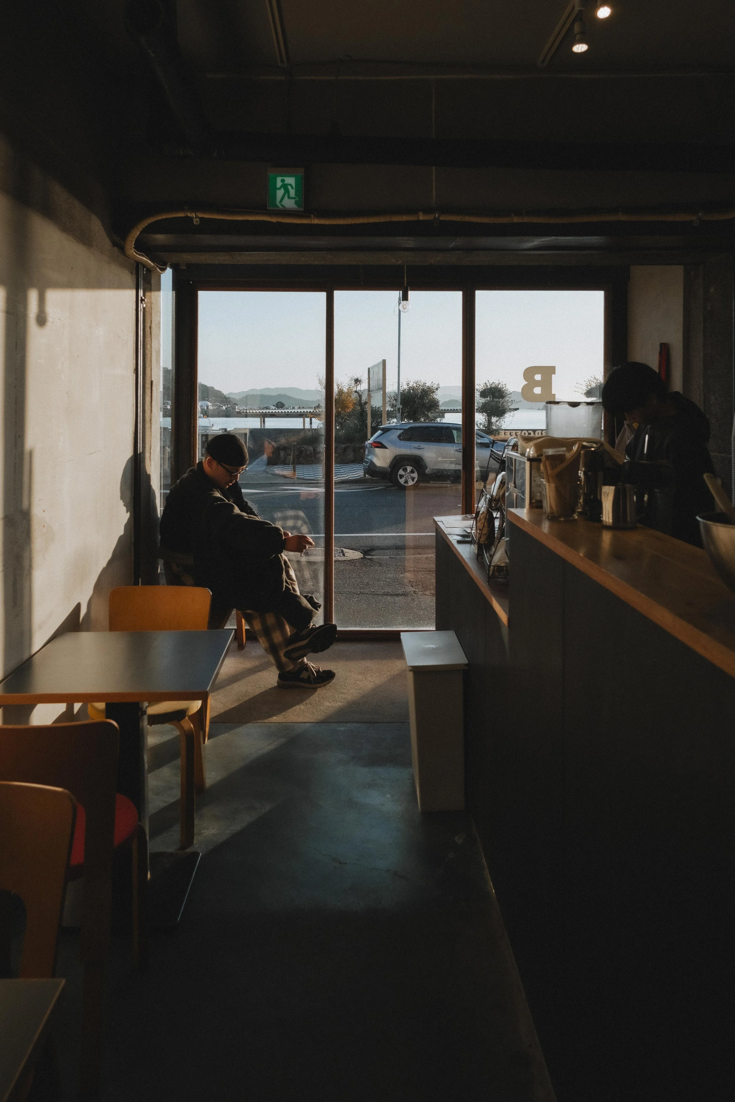 A cozy coffee shop interior with sunlight streaming through large front windows, showing parked cars outside, a person sitting near the window looking at their phone, and a barista working behind the counter.