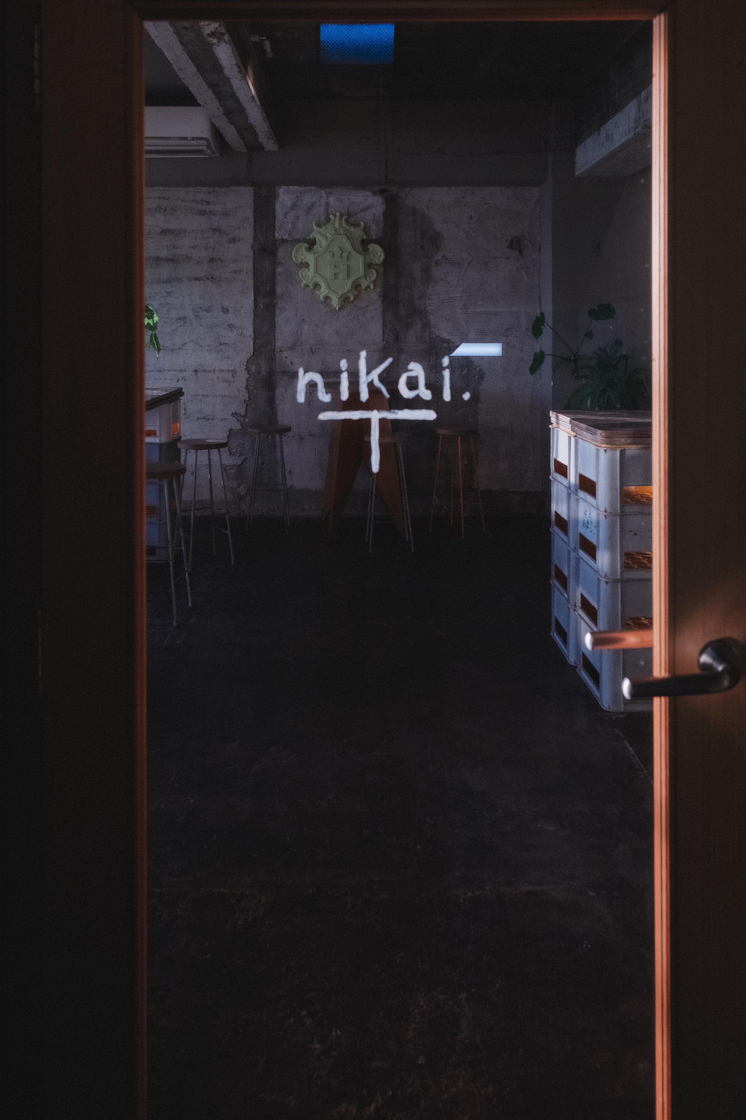 View through glass door into a dimly lit room with industrial decor, the word 'nikai.' written on the glass in white, and a green decorative sign on the wall.