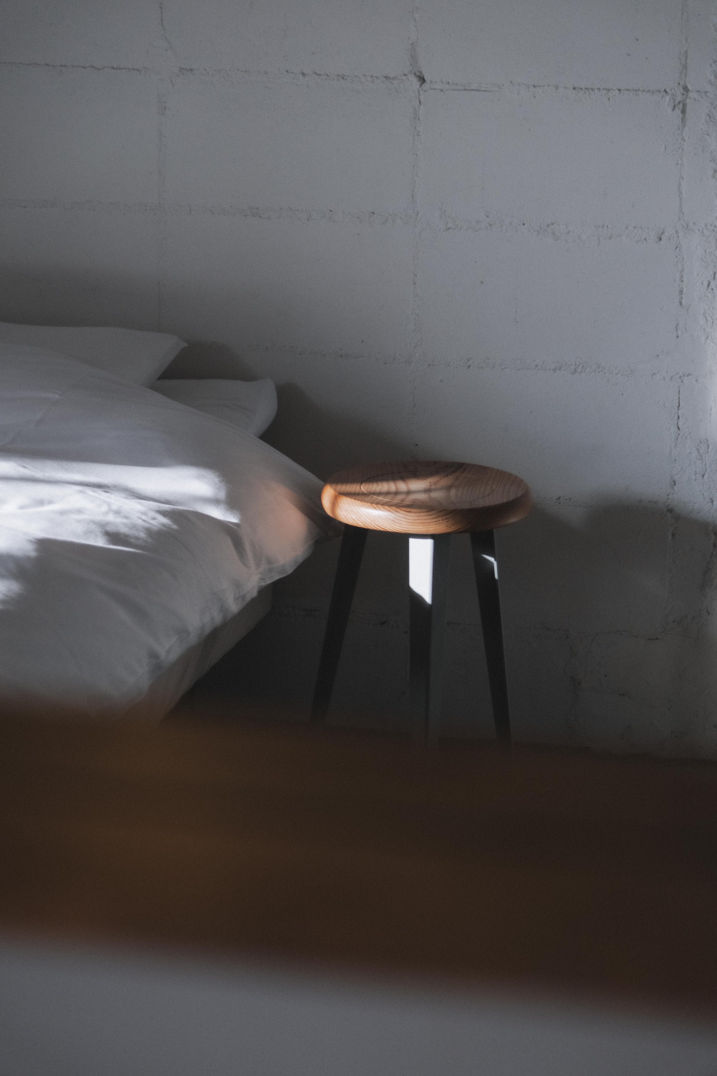 Close-up of a bed with white sheets and pillows next to a wooden stool with black legs against a textured white wall.