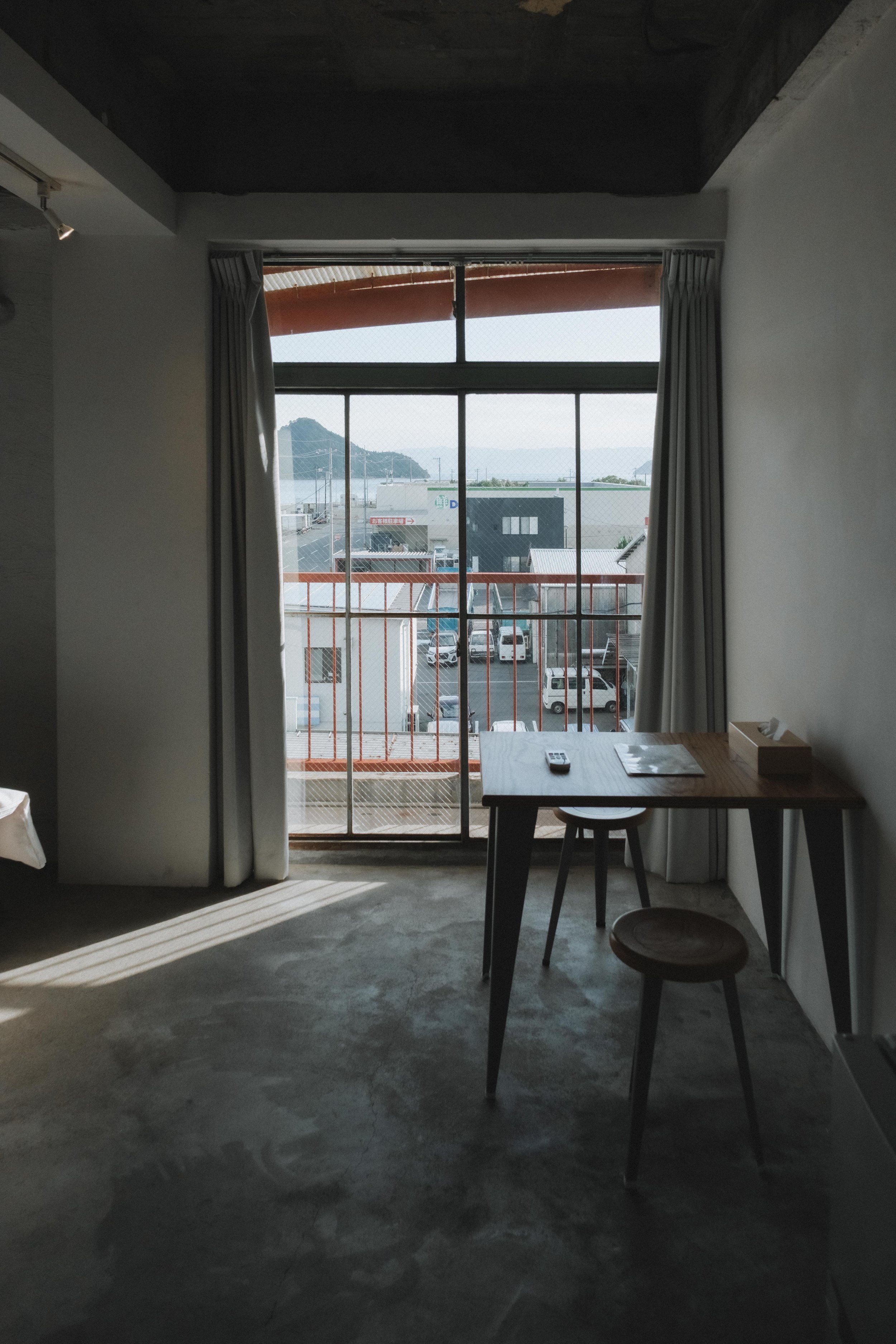 Interior of a room with a wooden desk, two stools, and a large window with curtains revealing an outdoor parking lot and some buildings.