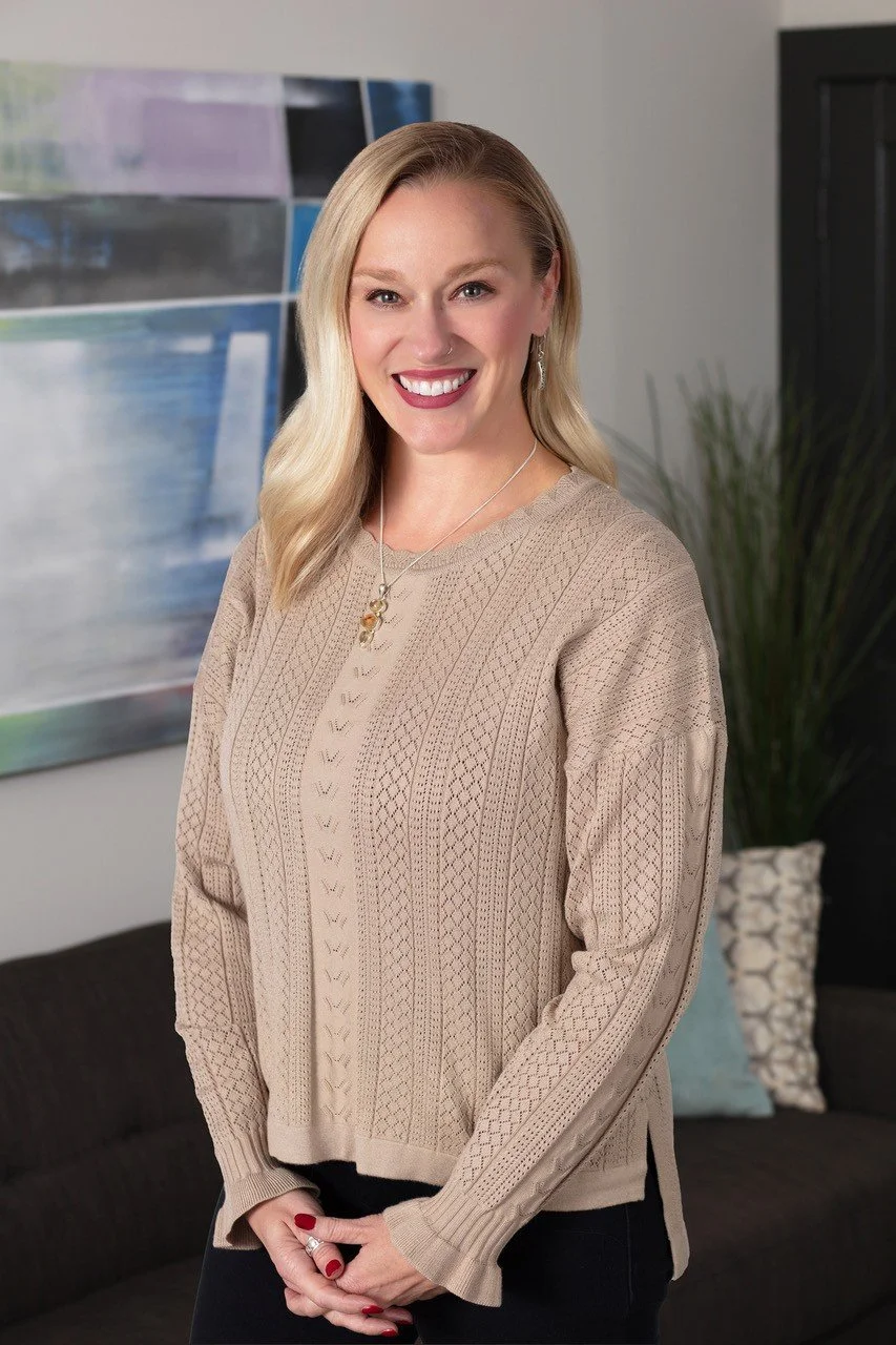 A woman with long blonde hair smiling, wearing a beige knit sweater and jewelry, standing in a living room with a black couch, colorful pillows, painting, and a tall green plant in the background.