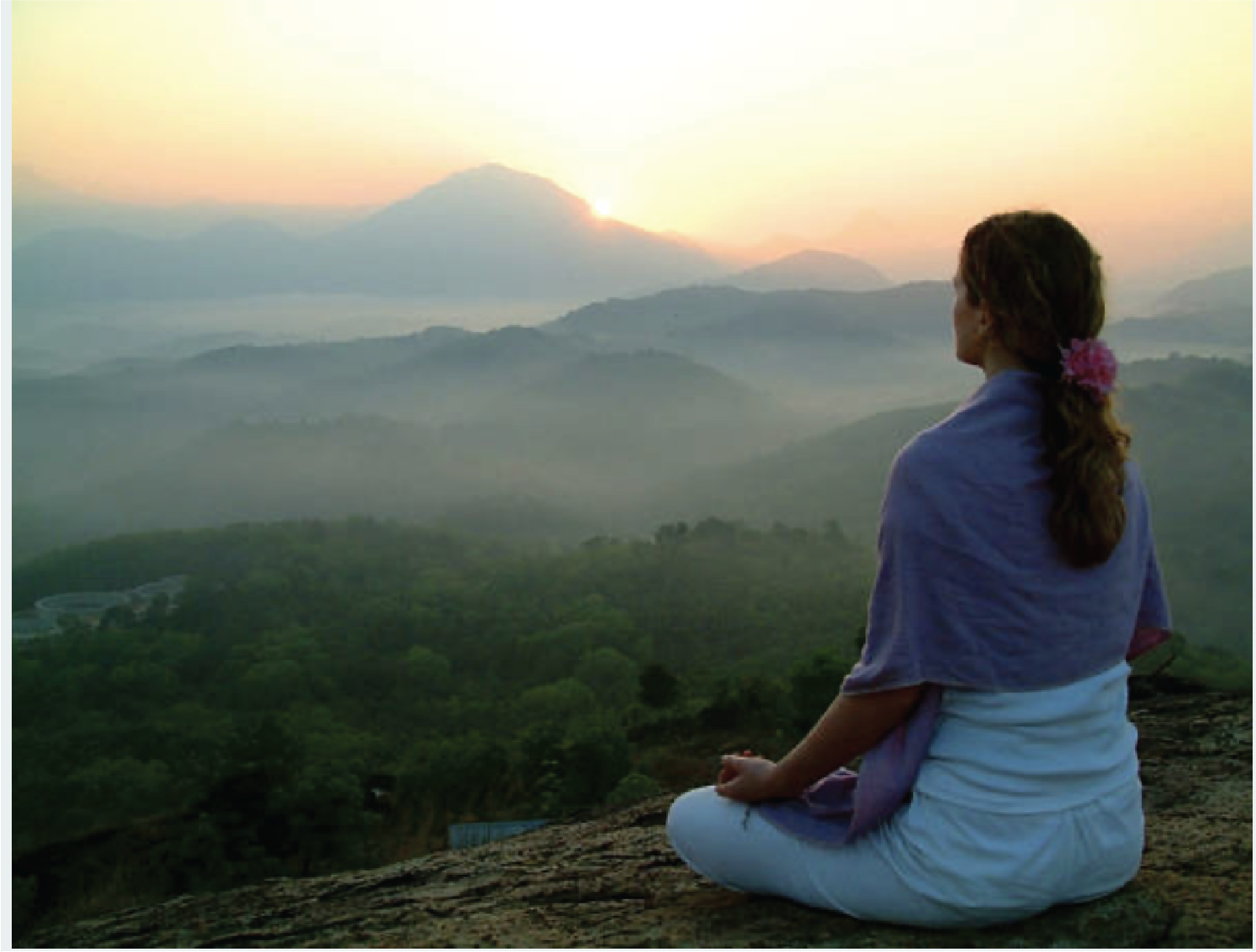 A woman sitting cross-legged on a rock, meditating or relaxing in front of a sunrise or sunset over a mountainous landscape with green forests and fog.