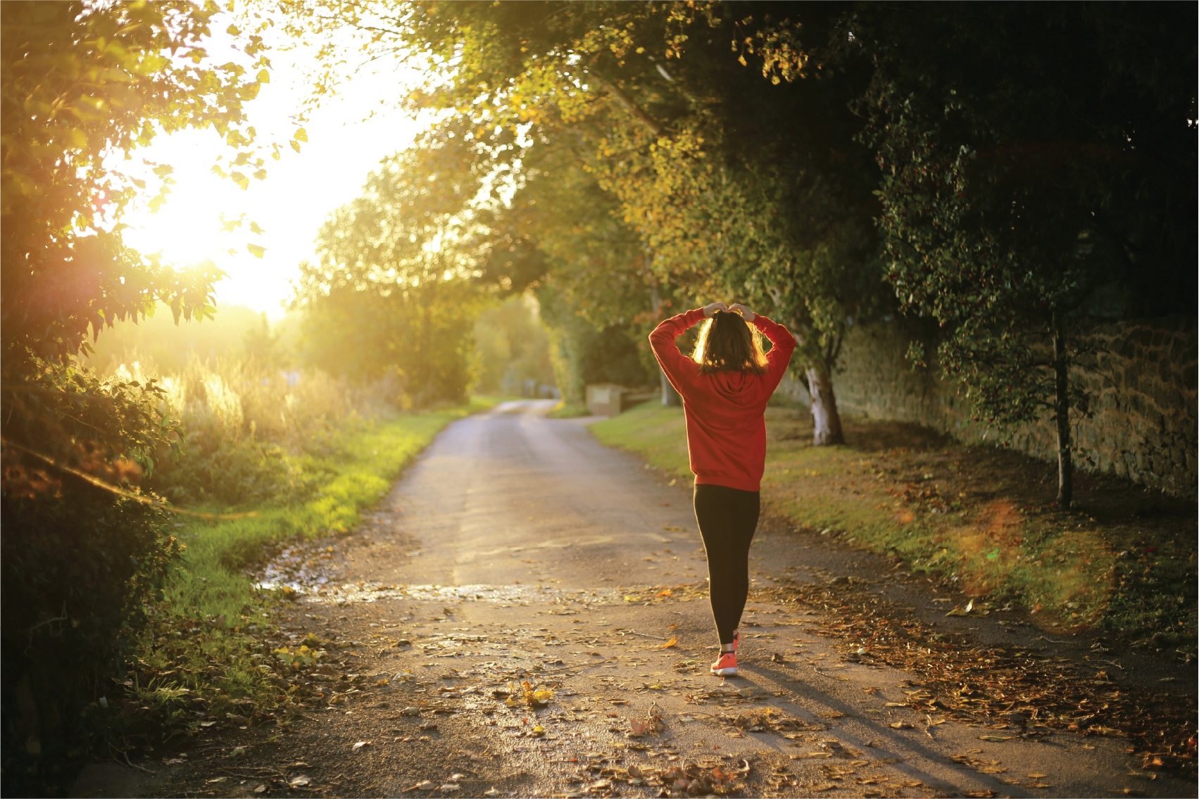 A woman in a red hoodie and black leggings walking along a wooded path at sunset, with her hands on her head.