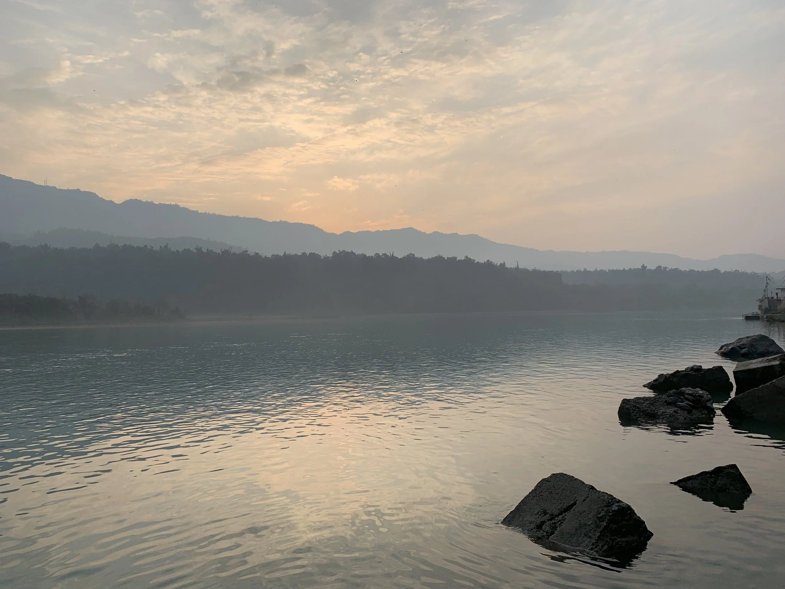 Calm river with rocks in the foreground, mountain ranges in the background, and a partly cloudy sky at sunset.