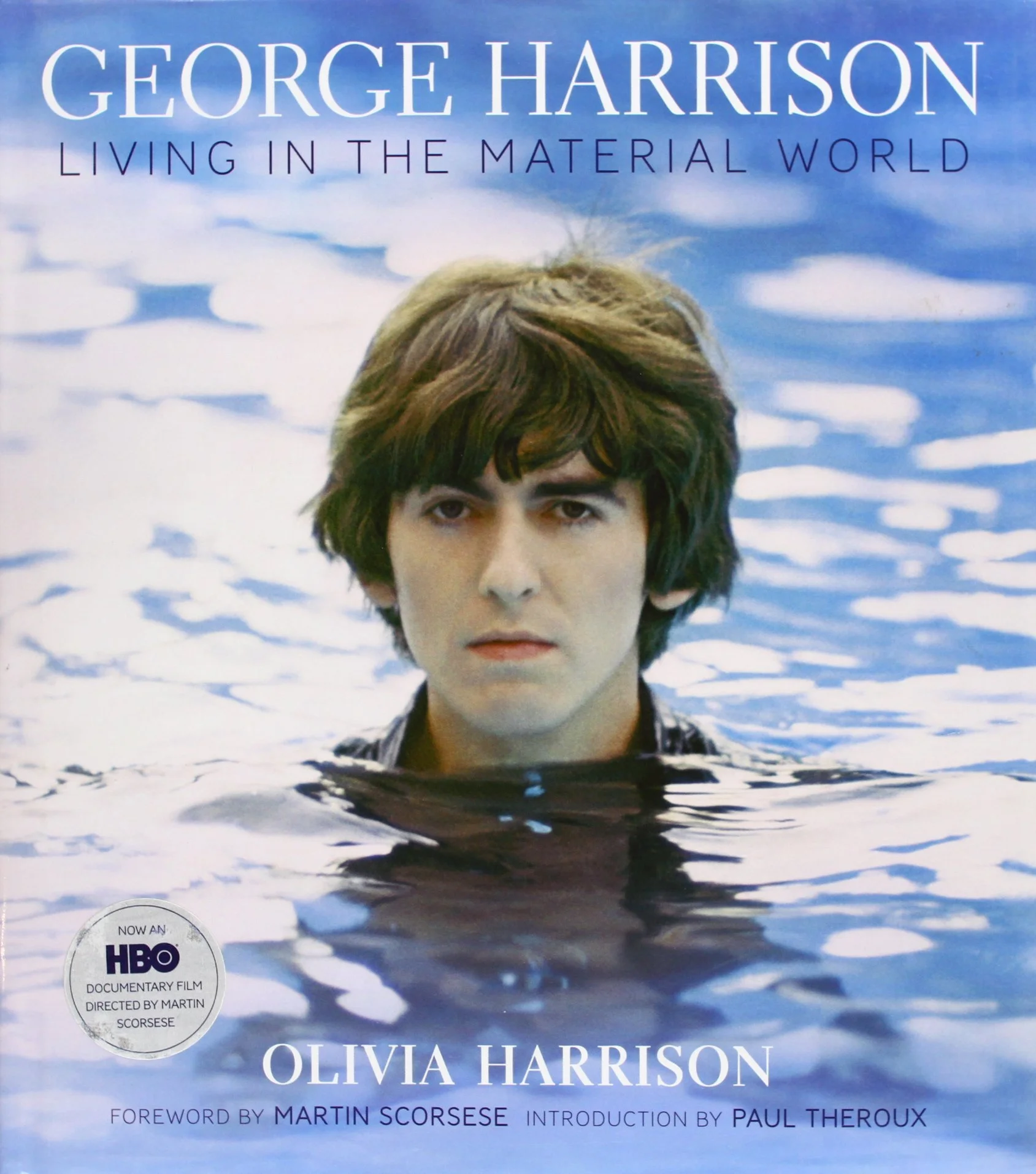 Book cover of George Harrison's "Living in the Material World" featuring a close-up of George Harrison's face, partially submerged in water with a blue sky background.