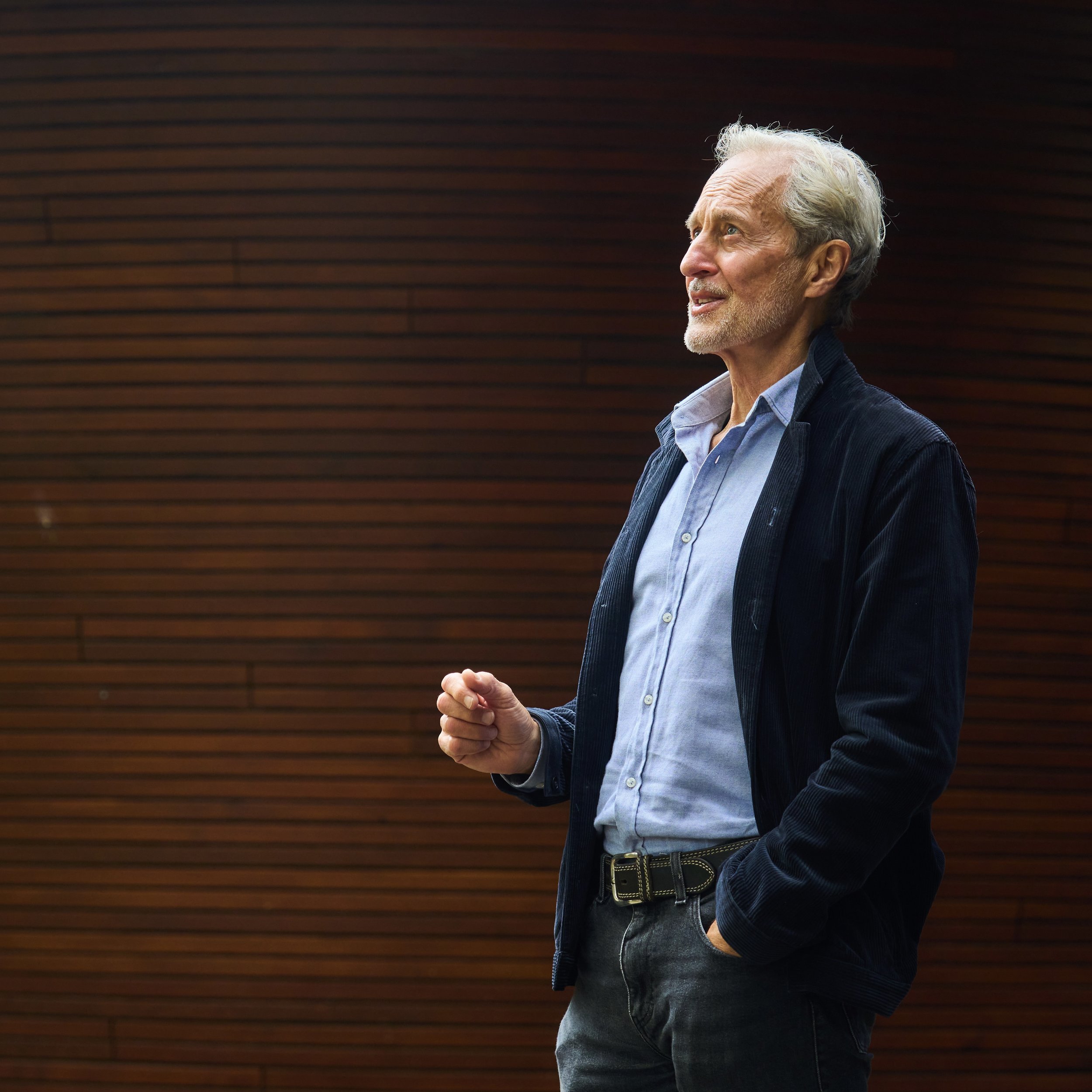 Anthony Thompson, an elderly man with gray hair and a beard standing against a wooden paneled wall, wearing a blue shirt and dark jacket, looking thoughtfully to the side.