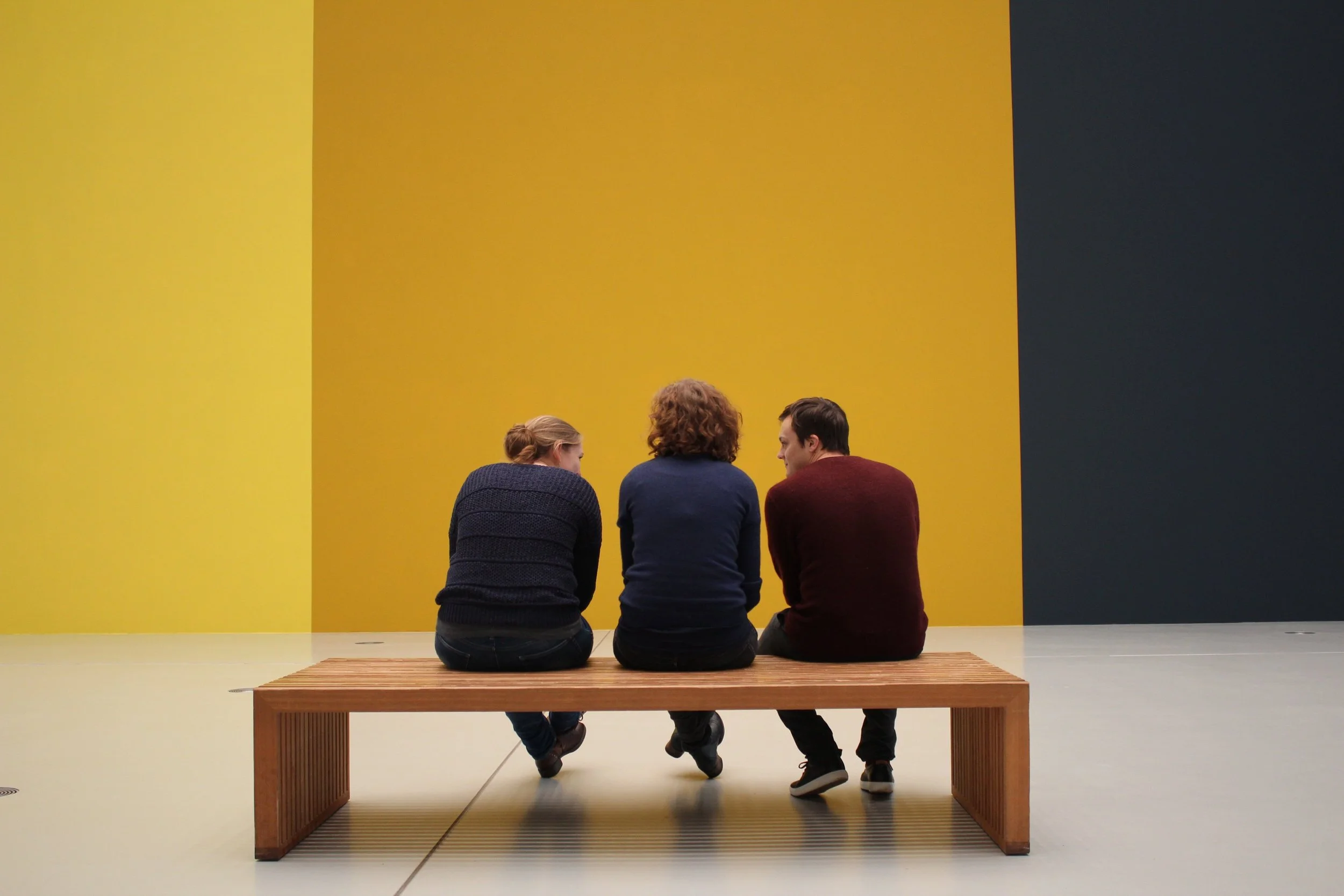 Three people sitting on a wooden bench in an art gallery, facing a large yellow and blue wall.