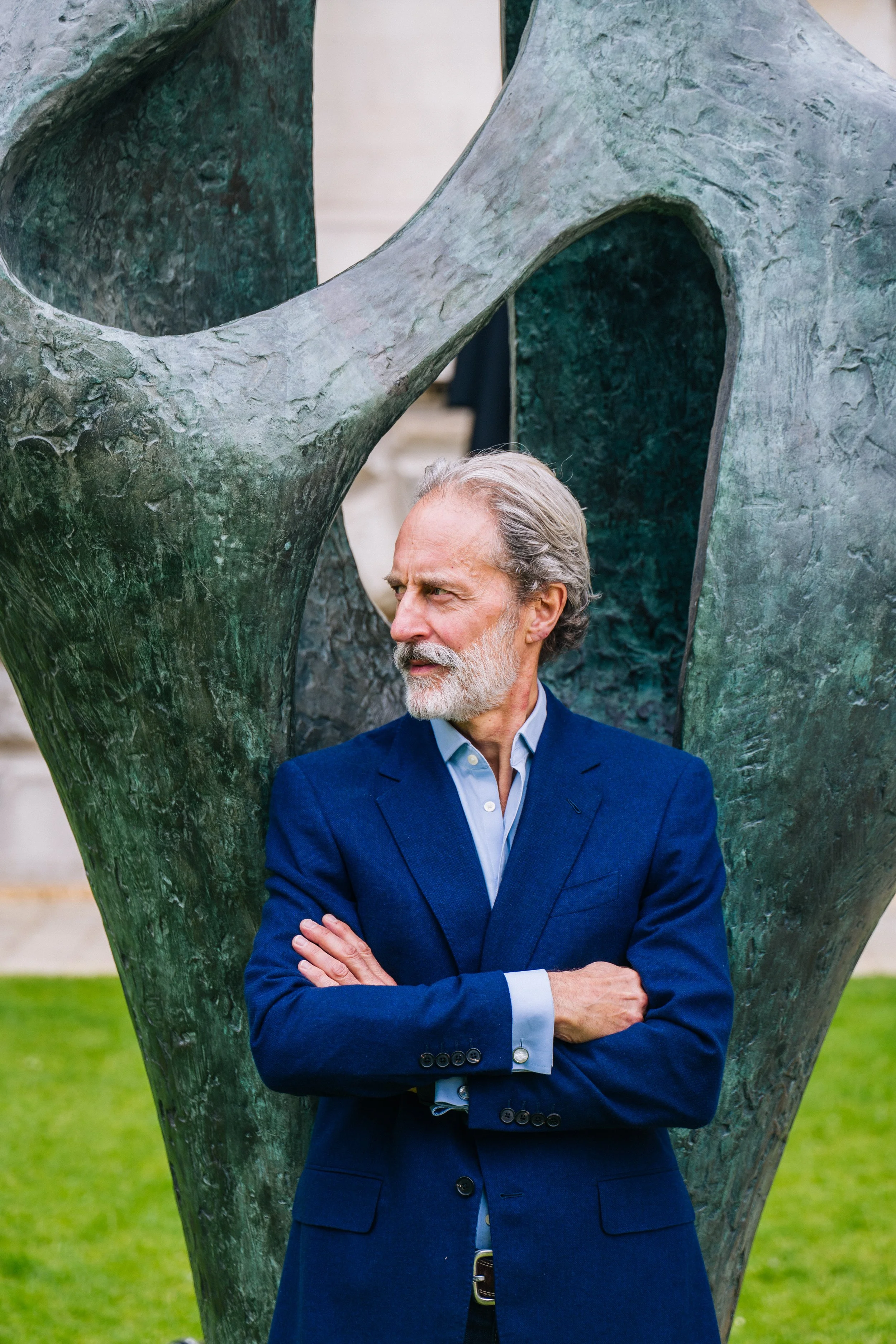 An older man with gray hair and a beard, Anthony Thompson, dressed in a blue suit with a light blue shirt, standing outdoors in front of an abstract metal sculpture, with arms crossed and a thoughtful expression.