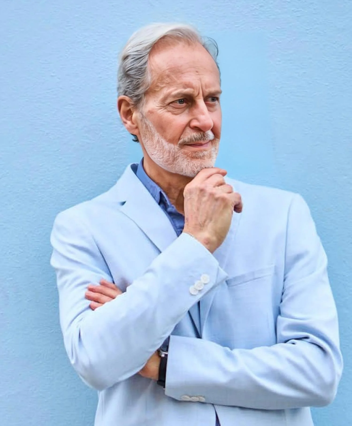 An elderly man with gray hair and a beard, Anthony Thompson, dressed in a light gray blazer and a blue shirt, standing in front of a light blue wall. He is resting his chin on his hand and looking thoughtfully into the distance.