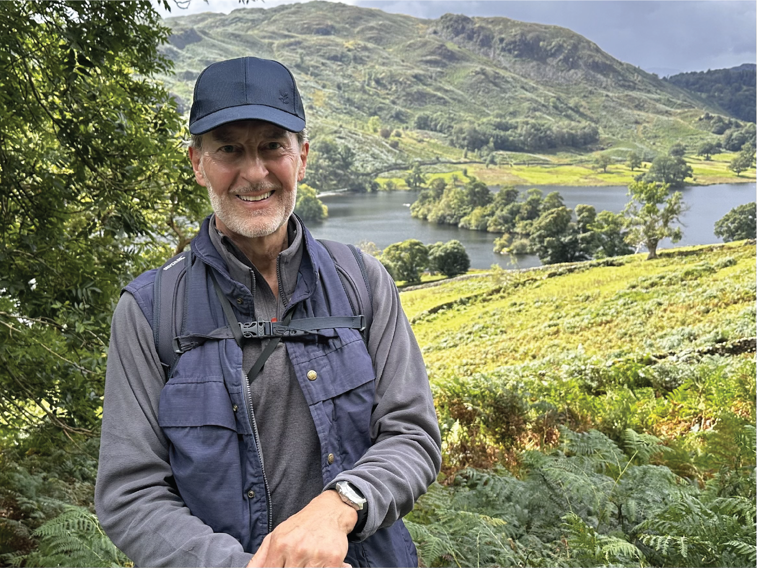 Anthony Thompson, a smiling man with a grey beard, wearing a blue cap, standing outdoors among green foliage with a scenic lake and hills in the background.