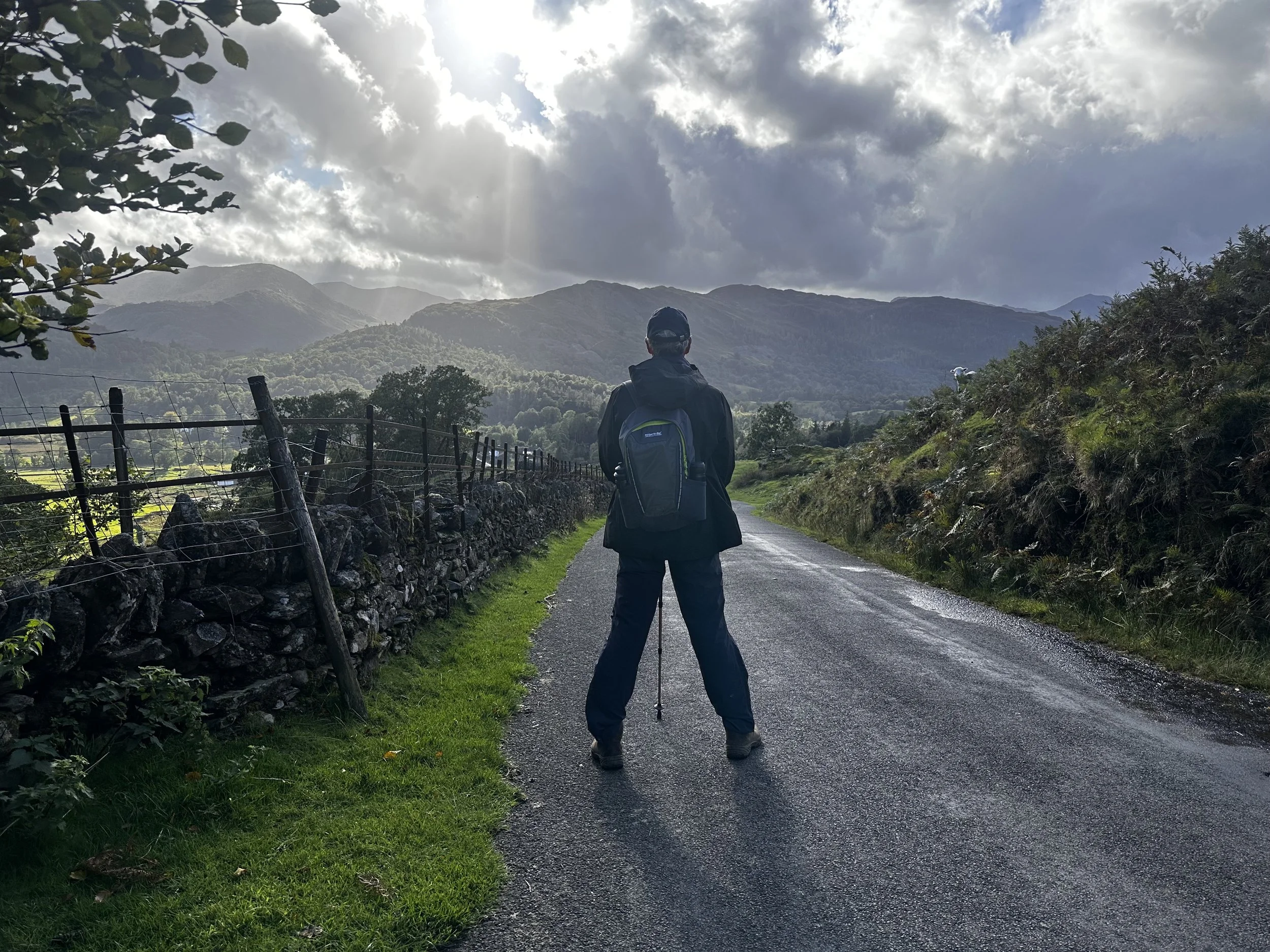 Person standing on a wet rural road with mountains in the background under cloudy sky.