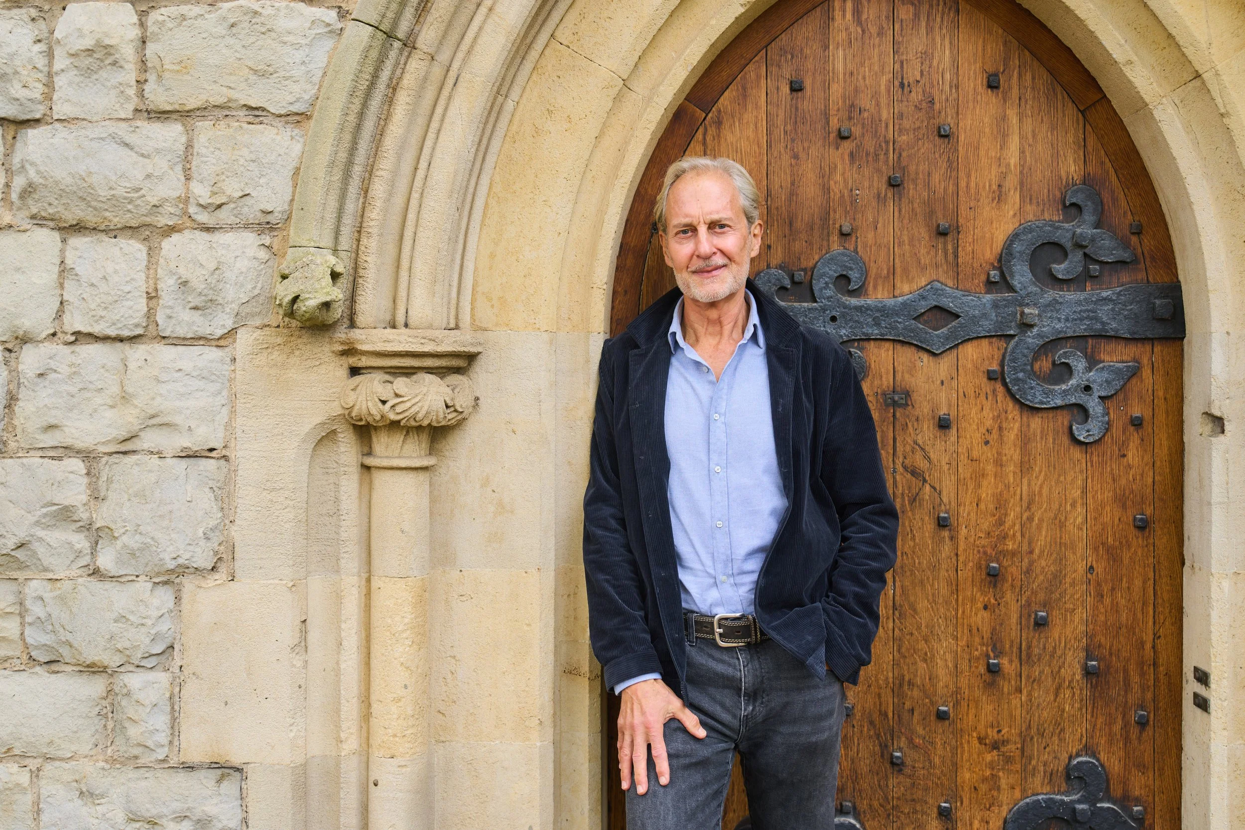 Anthony Thompson, an old man with gray hair and beard standing in front of a large wooden door with metal accents, part of a stone building exterior.