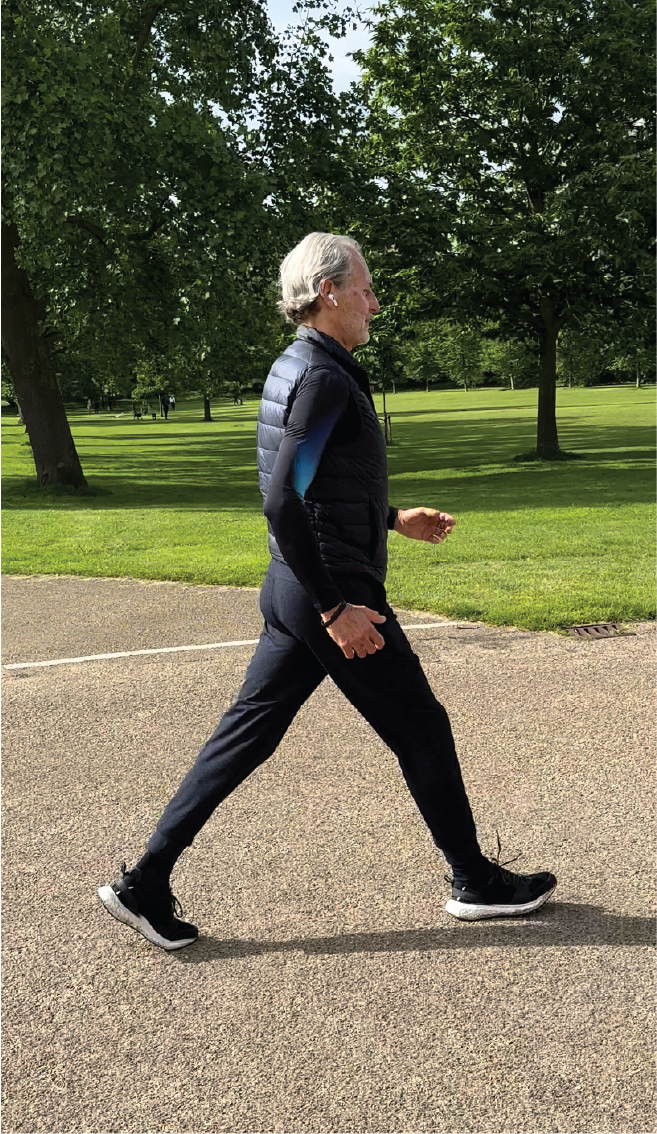 Anthony Thompson walking on a paved path in a park with green grass and trees in the background.