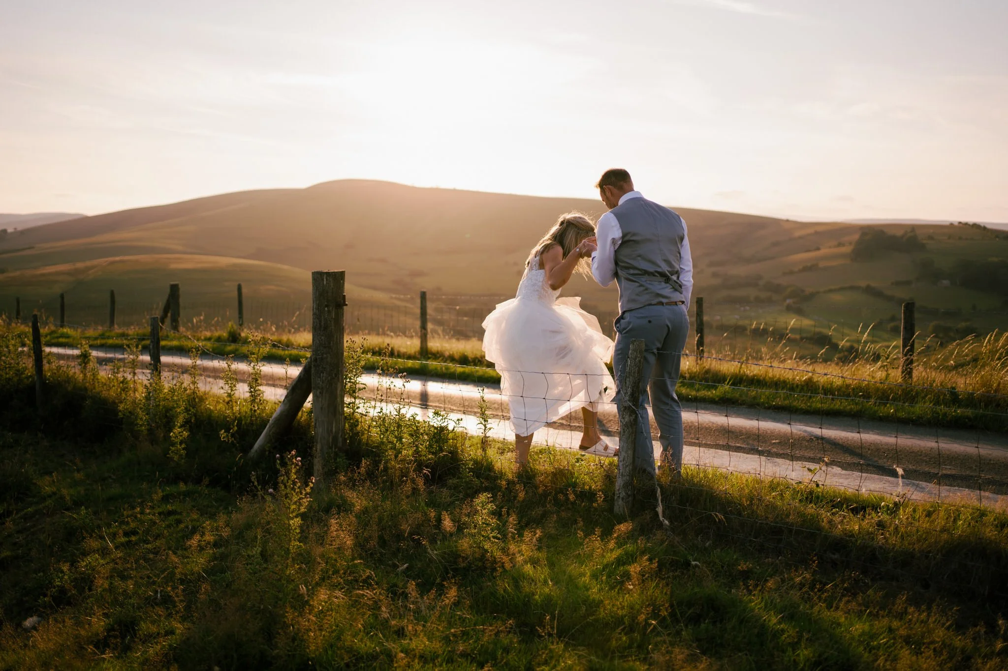 Groom helping bride cross a fence at sunset on a country road.