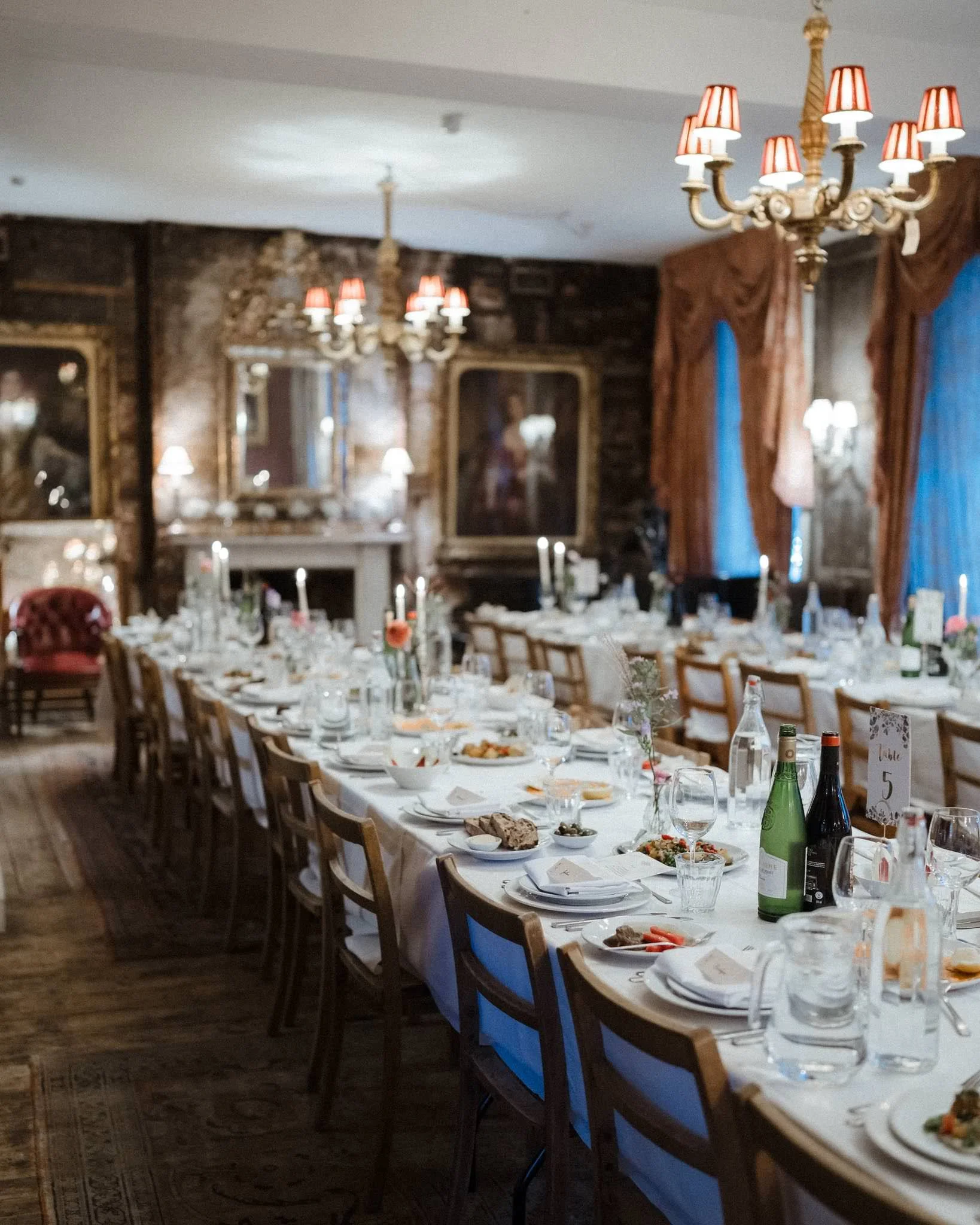 Elegant dining room set for a formal event with a long table, place settings, bottles of wine, and decorative centerpieces, in a richly decorated room with chandeliers, ornate mirrors, and draped curtains.