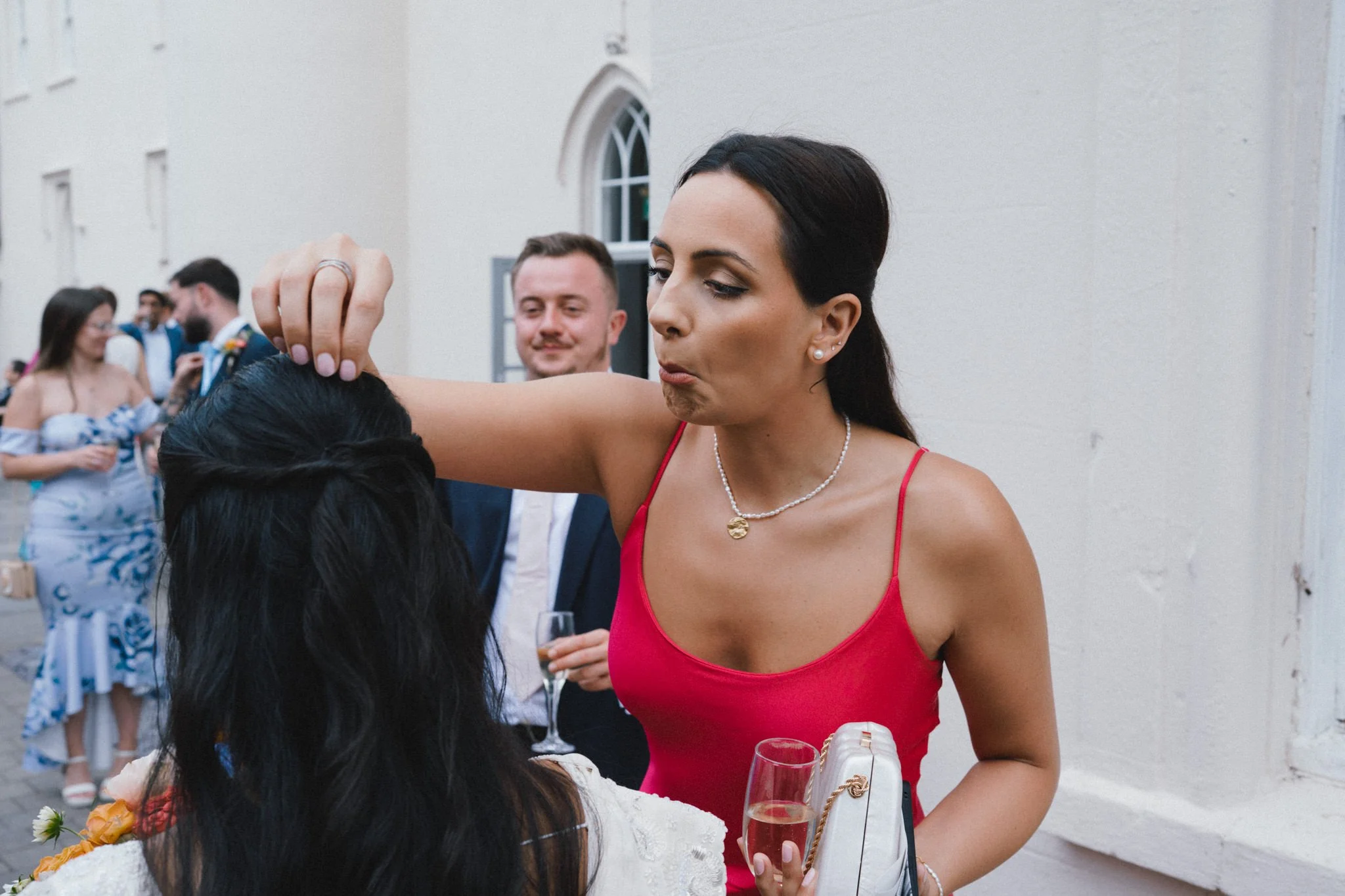 wedding photography of a female guest in a red dress, pulling confetti out of the bride's hair while pulling a funny face