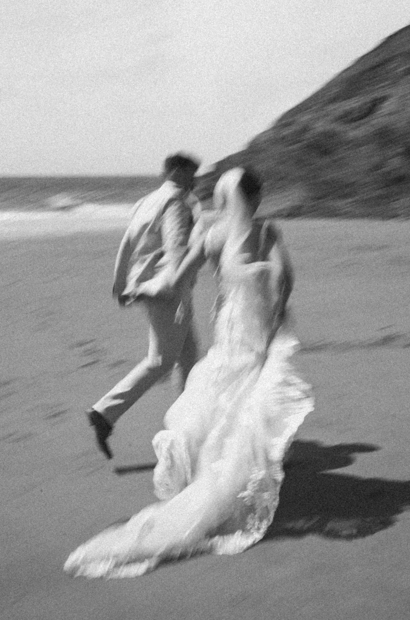 A slow shutter black and white photo of a bride and groom running down the beach at Mwnt in West Wales
