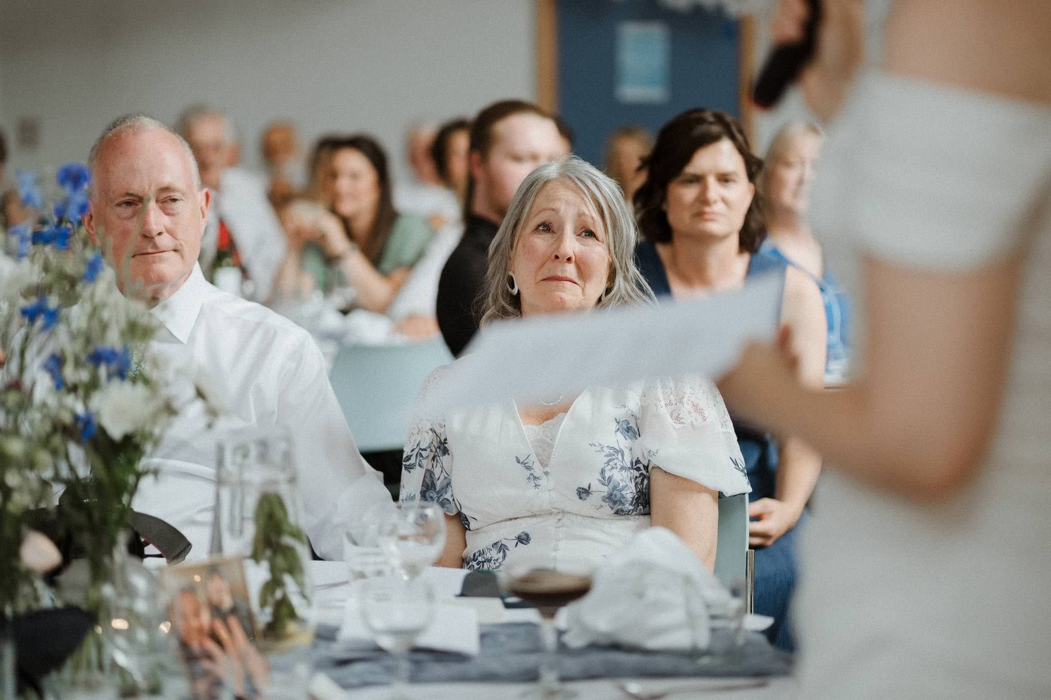 People attending a formal event, sit at a table with flowers, glasses, and photographs, looking at a speaker or presenter.