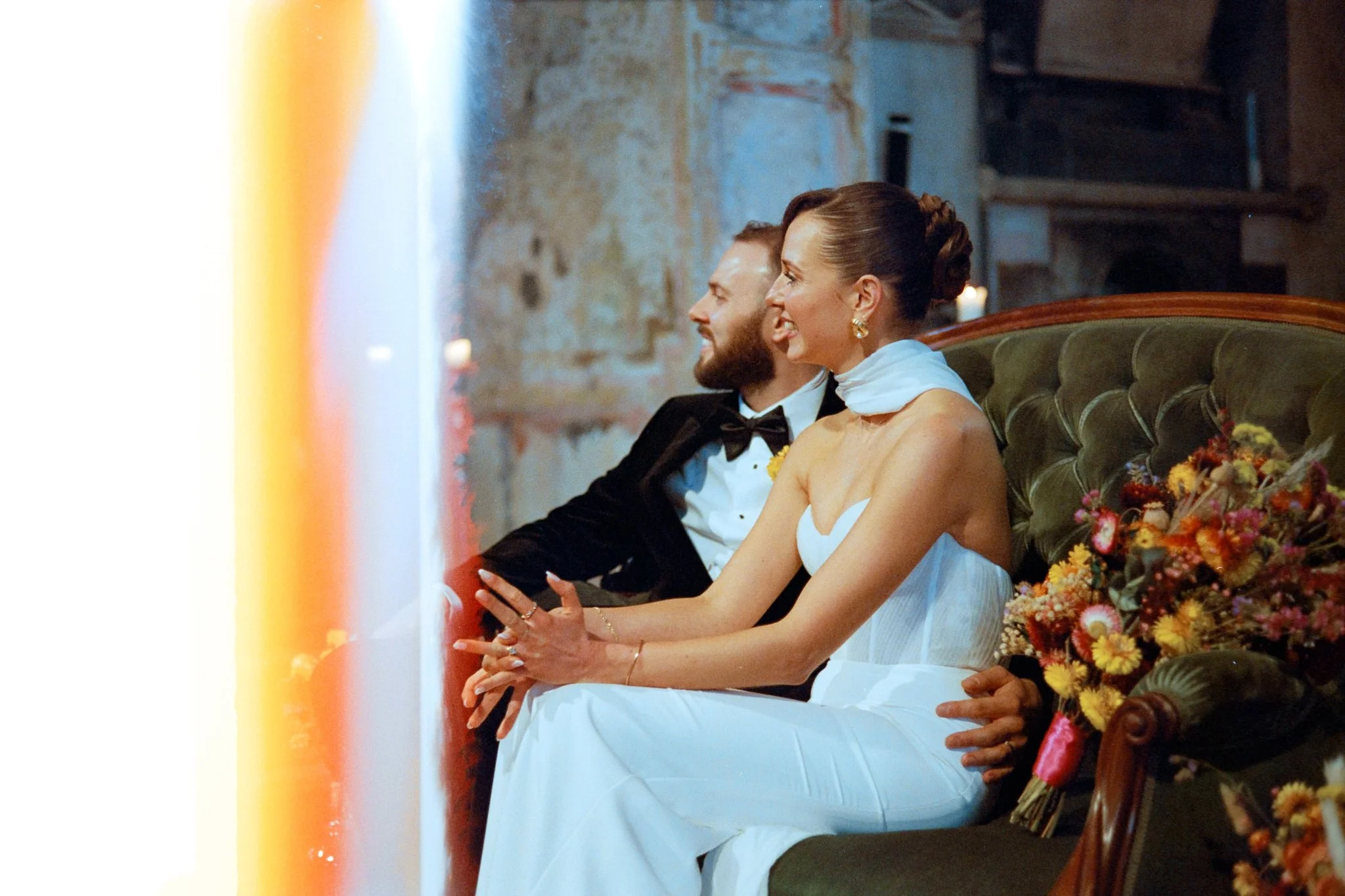 Bride and groom sitting side by side in a wedding ceremony, smiling, with colorful flowers in front of them and candles in the background.